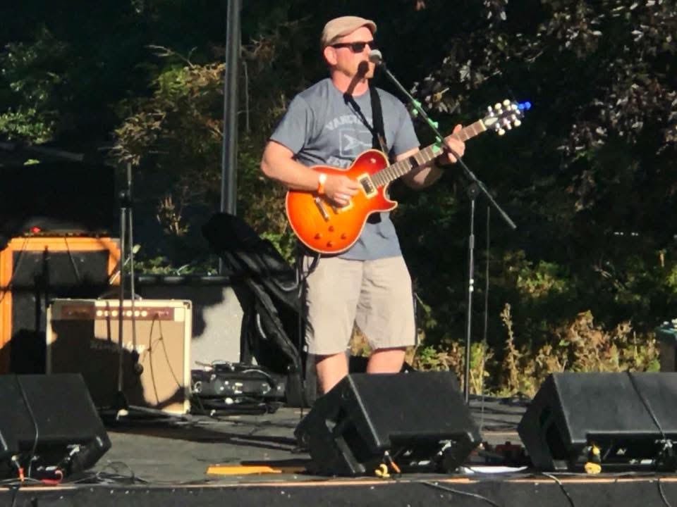 A man playing an orange electric guitar on an outdoor stage, wearing sunglasses, a hat, a gray T-shirt, and khaki shorts.
