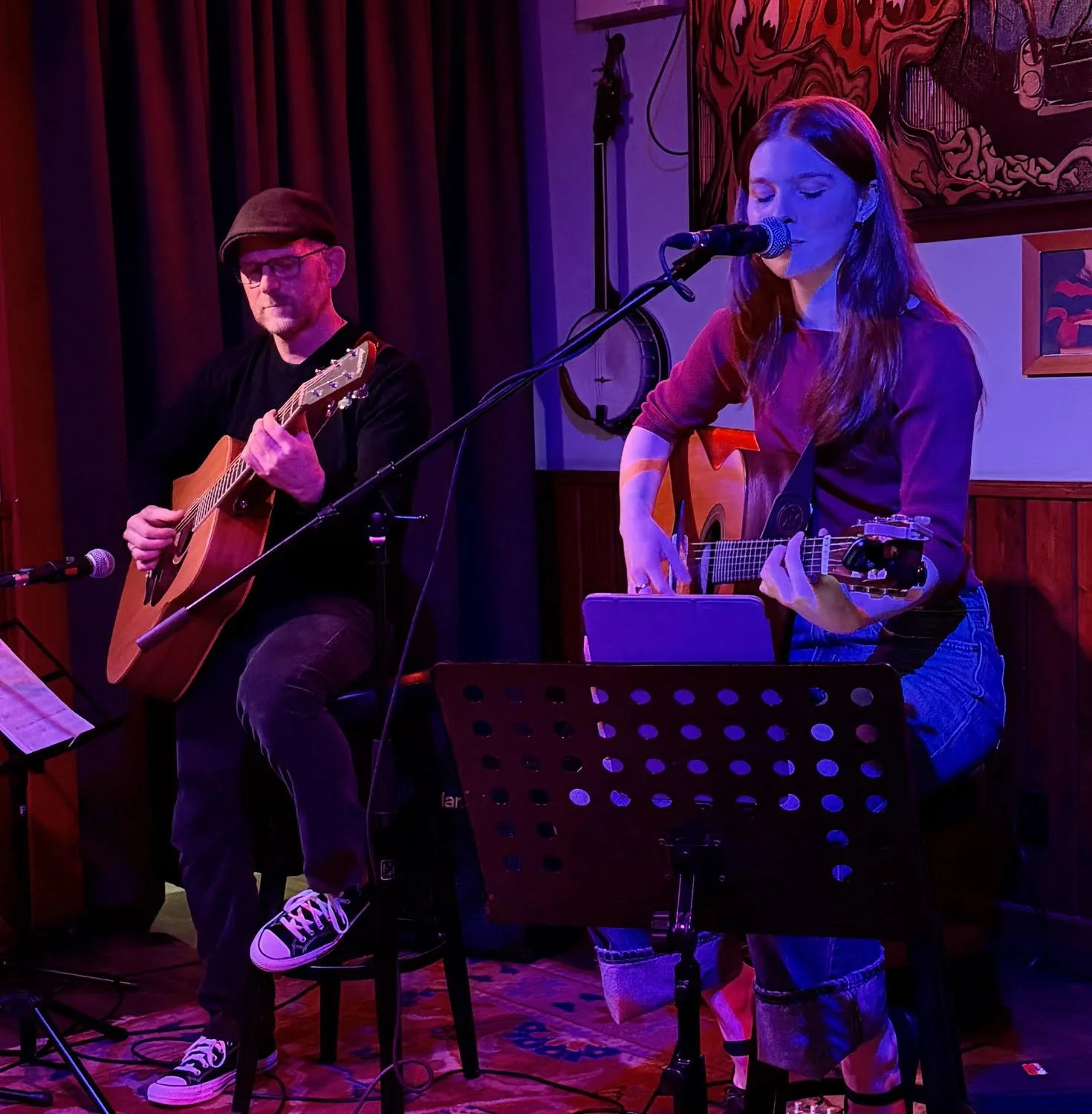 A man and woman performing musical acoustic set, the woman singing into a microphone and the man playing guitar at an indoor venue with warm lighting.