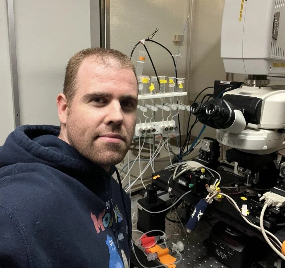 A man in a dark hoodie sits in a laboratory with scientific equipment, including a microscope and tubes connected by wires, around him.