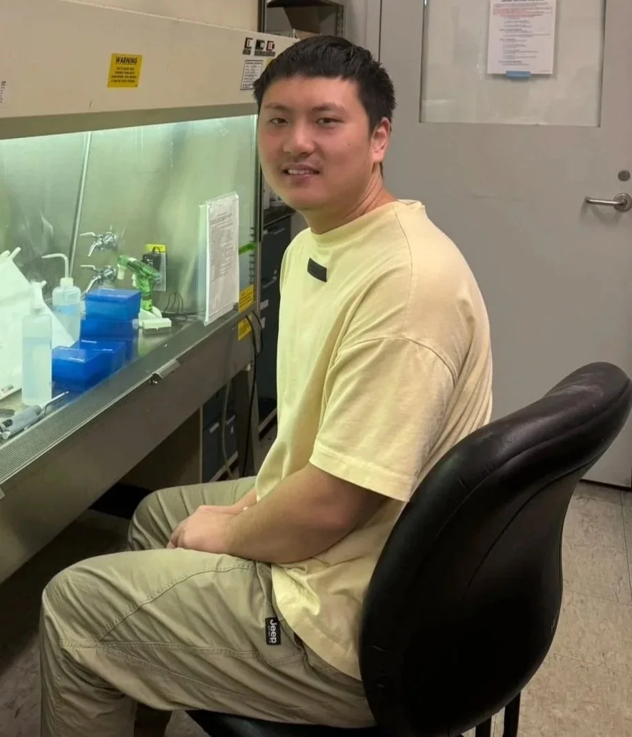 A young man sitting on a black chair in a laboratory or medical setting, with lab equipment and supplies on a workbench beside him.