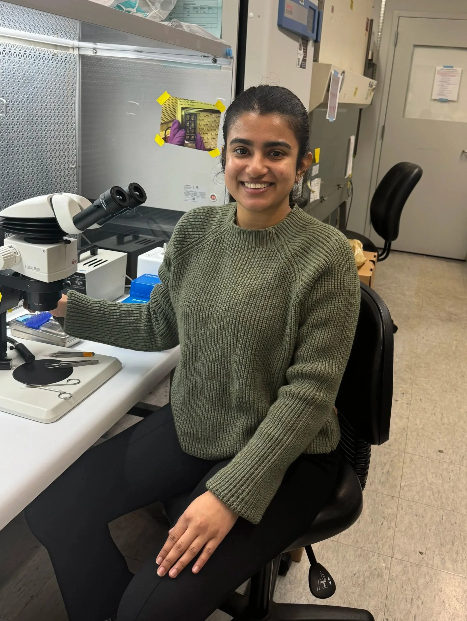 A woman sitting at a laboratory workbench with a microscope and laboratory tools, smiling at the camera. She is wearing a green sweater and black pants, with her right hand resting on her lap. The lab has equipment and cabinets in the background.