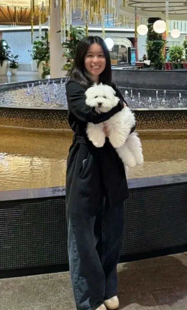A woman in black clothing holding a fluffy white dog inside a mall or hotel lobby with decorative lighting, plants, and a fountain in the background.