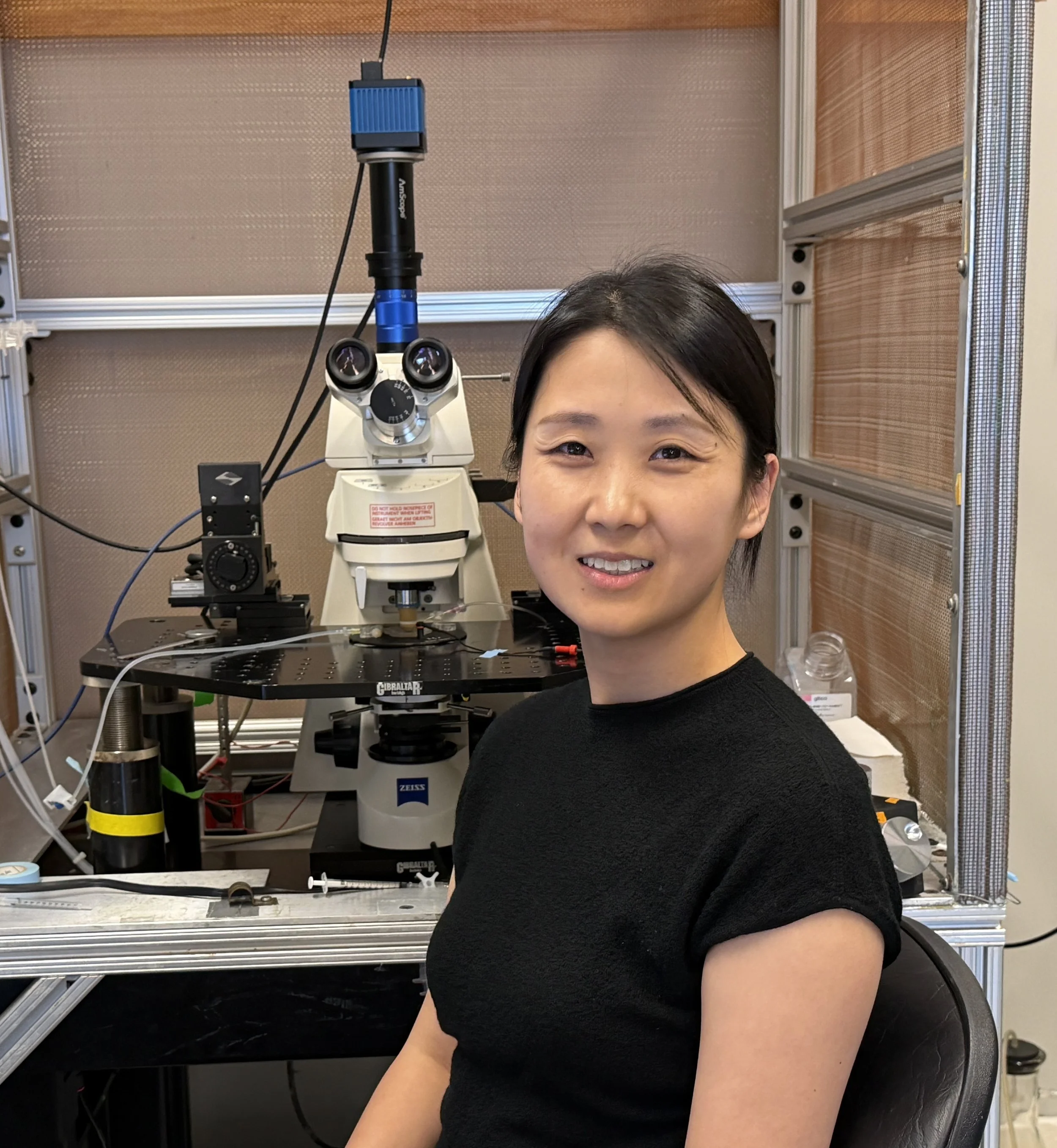 A woman in a black shirt standing next to a microscope in a laboratory setting.