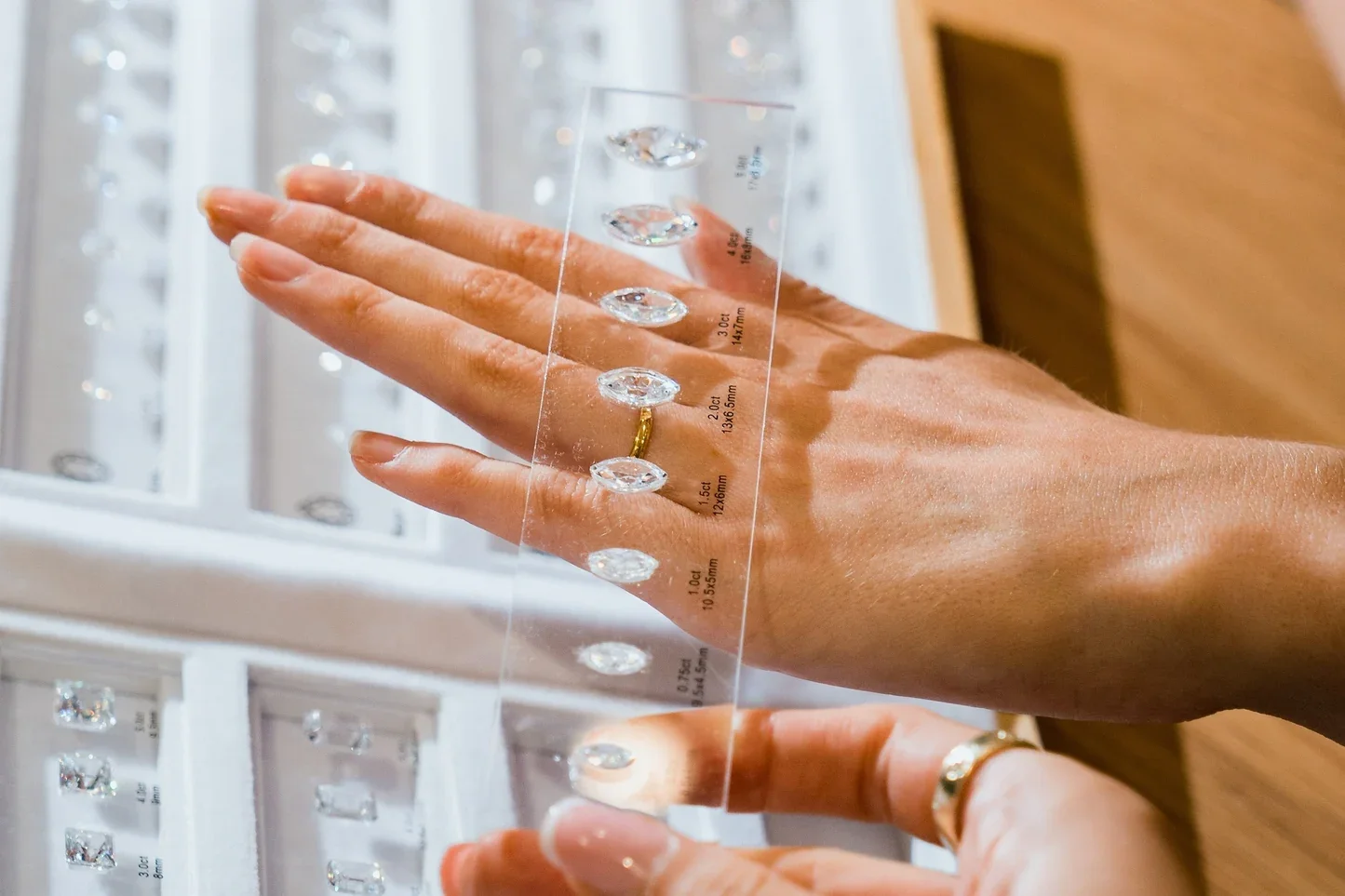 A hand with a gold ring on the ring finger holds a transparent acrylic gauge with various cut diamonds and gemstone measurements. In the background, display cases filled with more jewelry are visible.