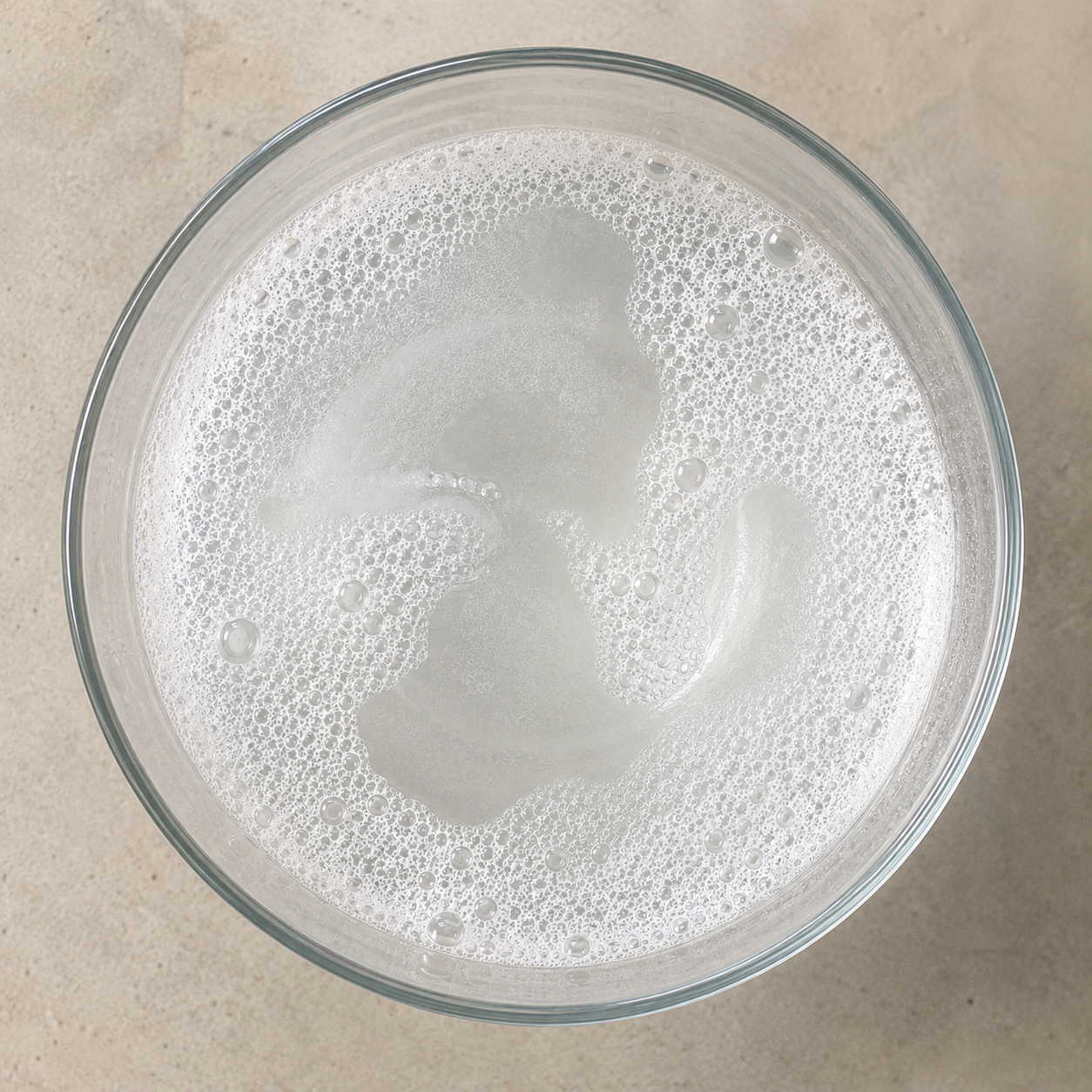 A clear glass of water with bubbles on top, viewed from above, on a beige surface.