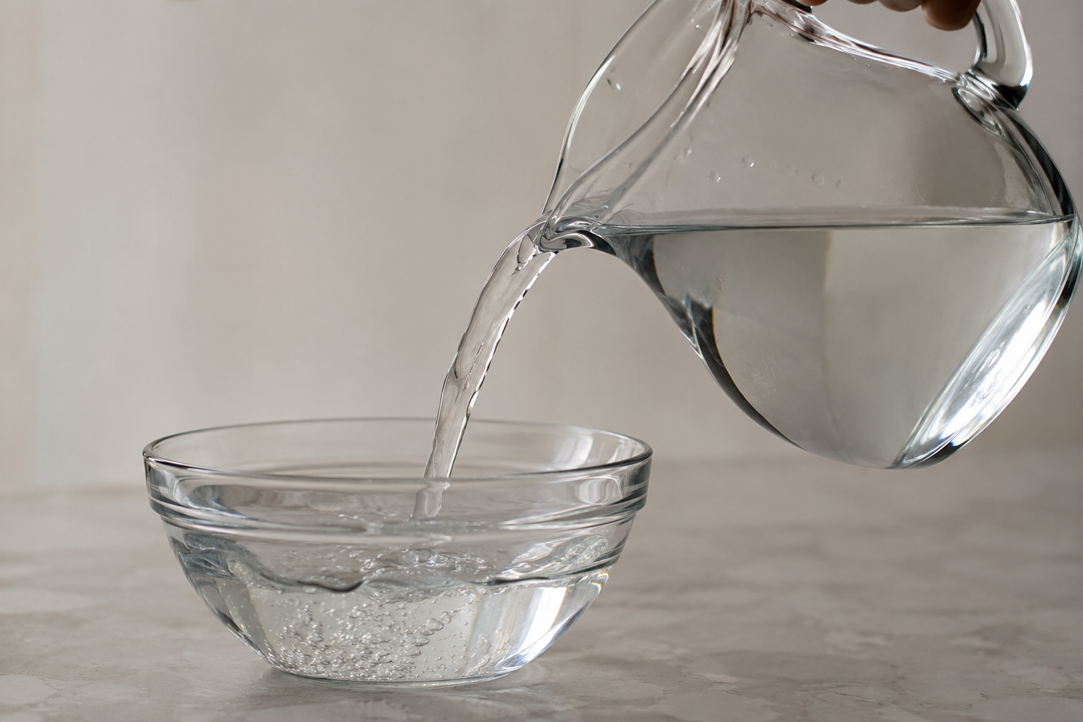 Close-up of a glass pitcher pouring water into a glass bowl on a light-colored surface.