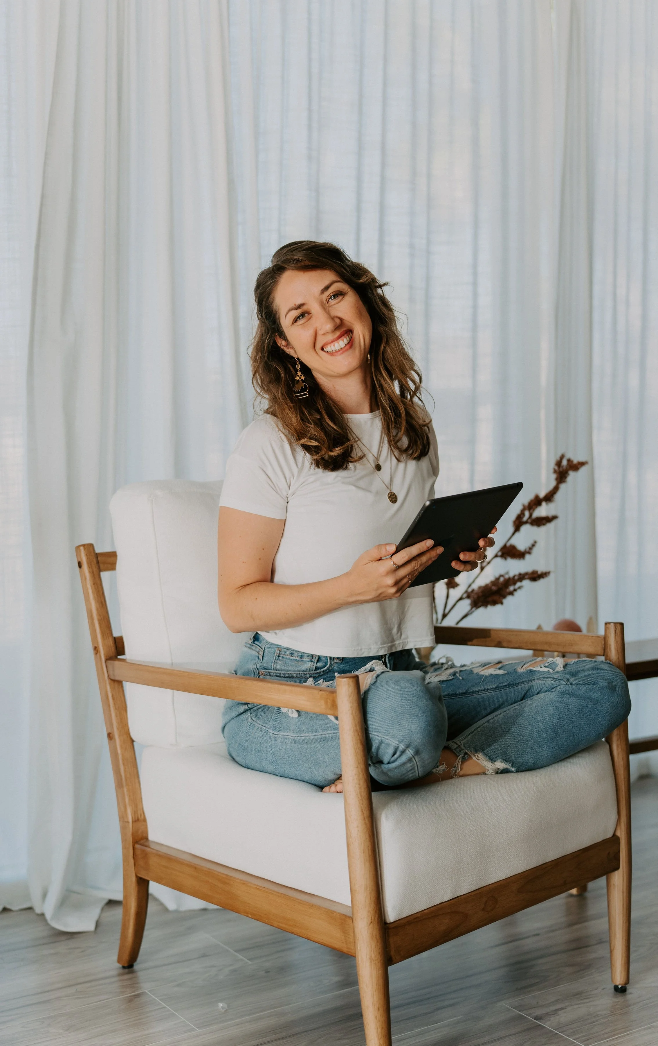 A young woman with wavy brown hair, wearing a white t-shirt and ripped jeans, sitting cross-legged on a wooden armchair with a white cushion, holding a black tablet, smiling, as she reads creative writing competition entries.