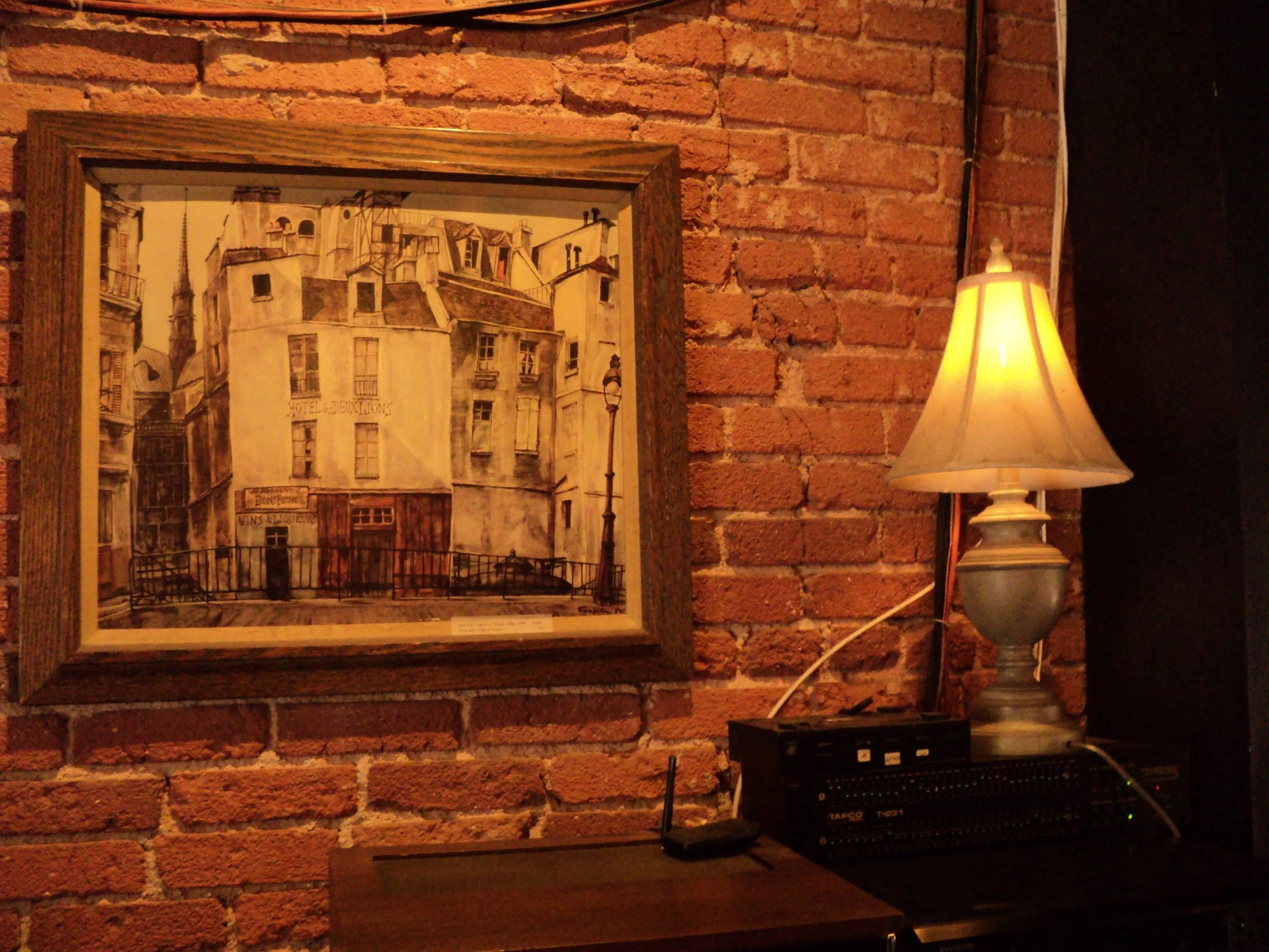 Decorative framed drawing of Parisian buildings on a brick wall, illuminated by a table lamp with a beige lampshade.