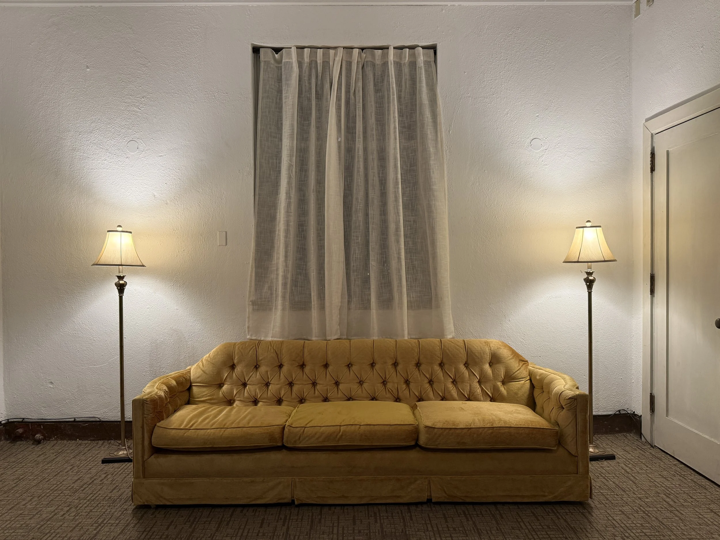 A living room with a yellow velvety tufted sofa, two floor lamps on either side, a window with sheer curtains, a white textured wall, and a closed door to the right.