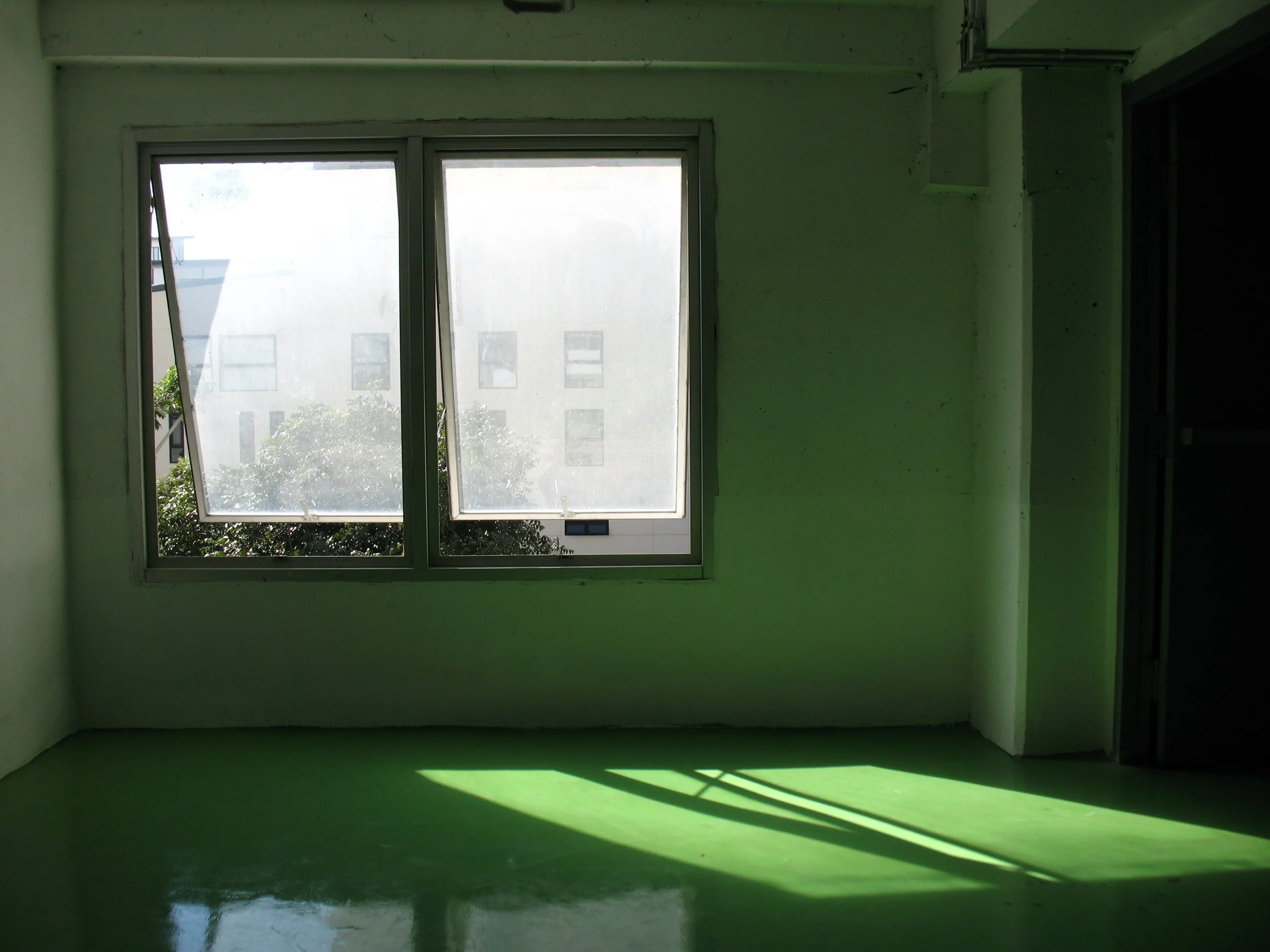 Empty room with green walls, large open window, sunlight casting shadows on the floor, and a view of neighboring buildings and trees outside.