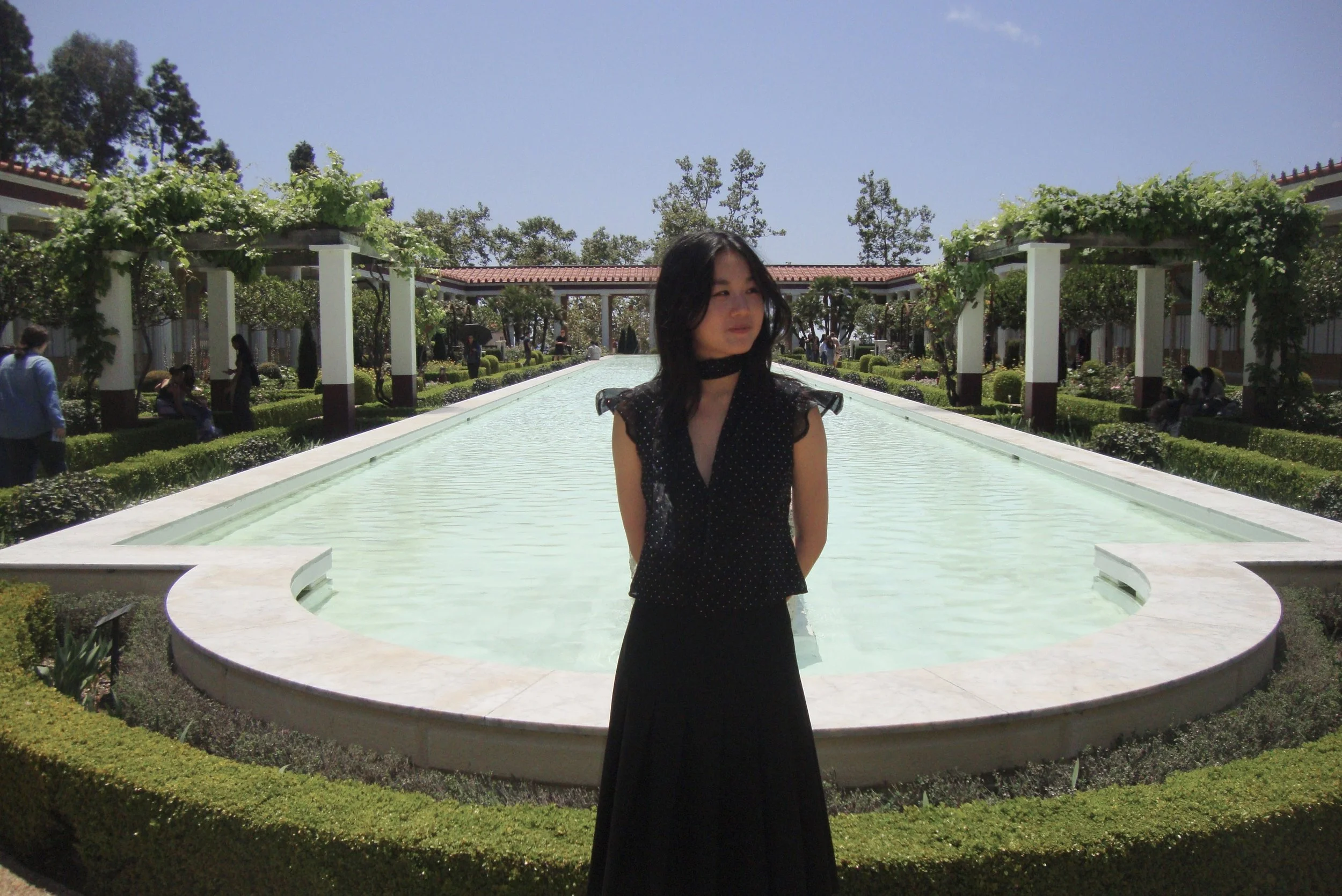 A woman with long black hair wearing a black dress with polka dots stands in front of a rectangular water fountain in a landscaped garden with trees, shrubs, and walkways, under a blue sky with few clouds.