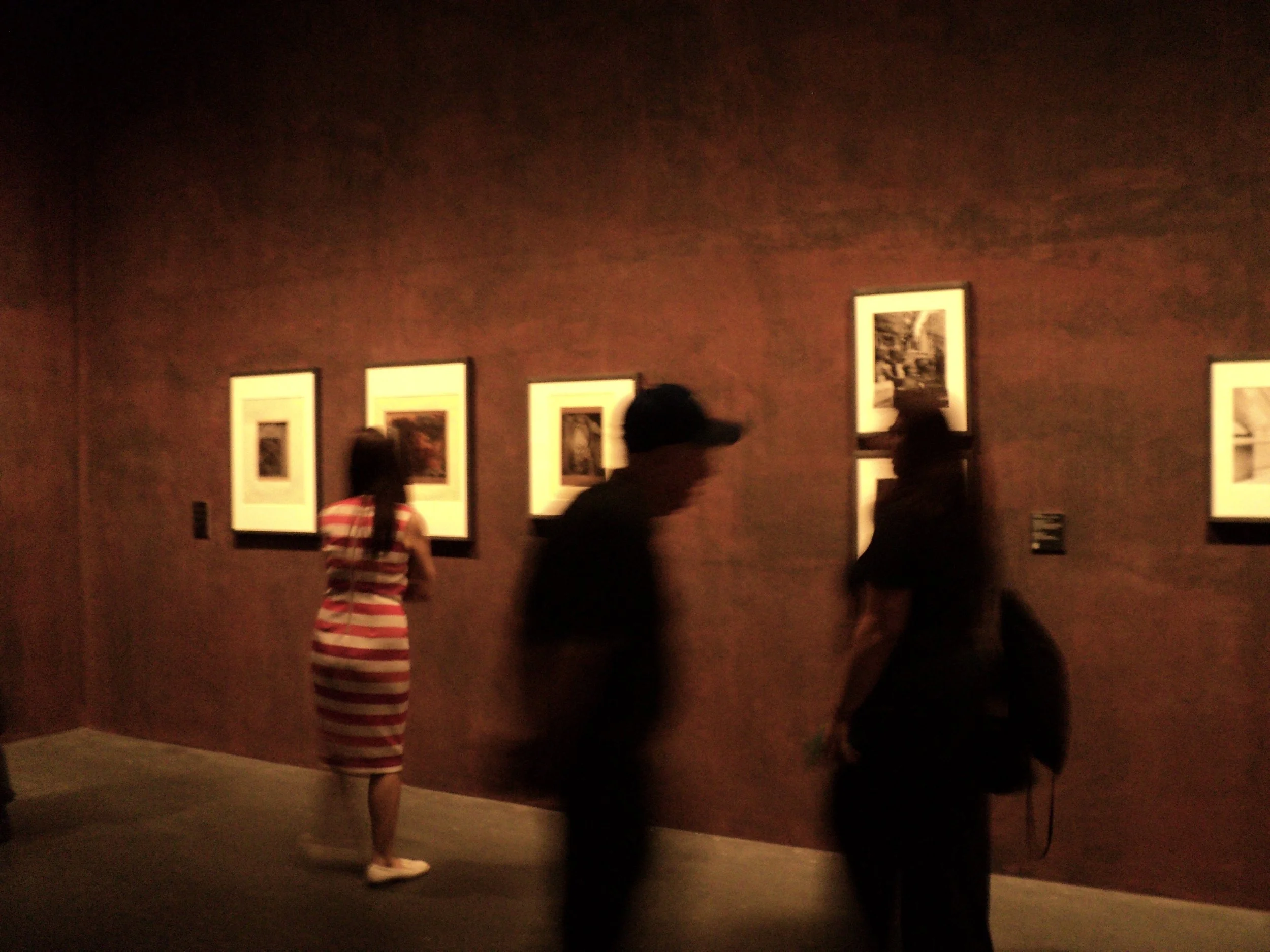 People viewing framed artwork in a gallery with brown walls and framed pictures.