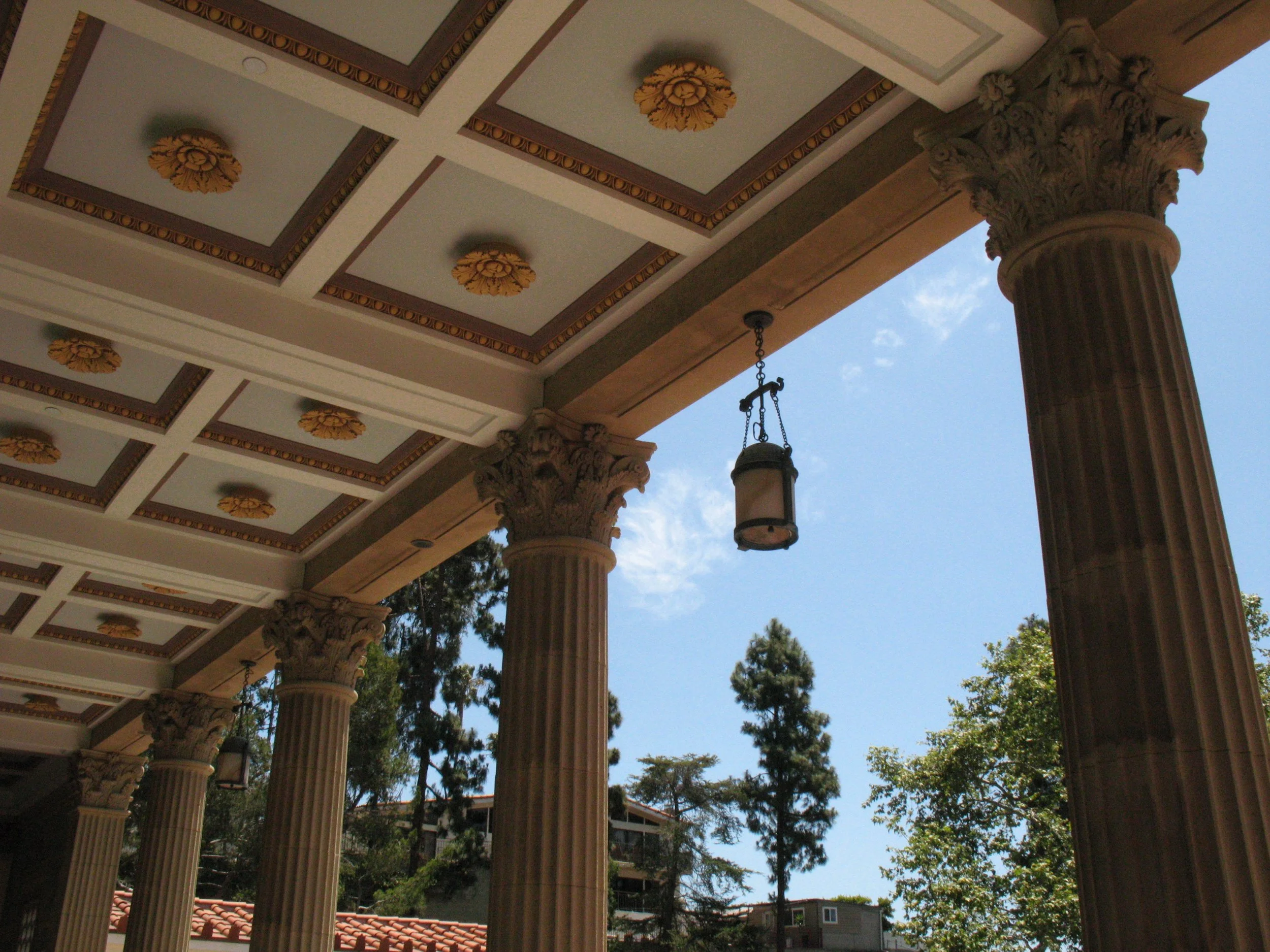 Exterior view of a classical building with tall Corinthian columns, decorated ceiling with gold accents, and hanging lanterns, framed by trees with a blue sky in the background.