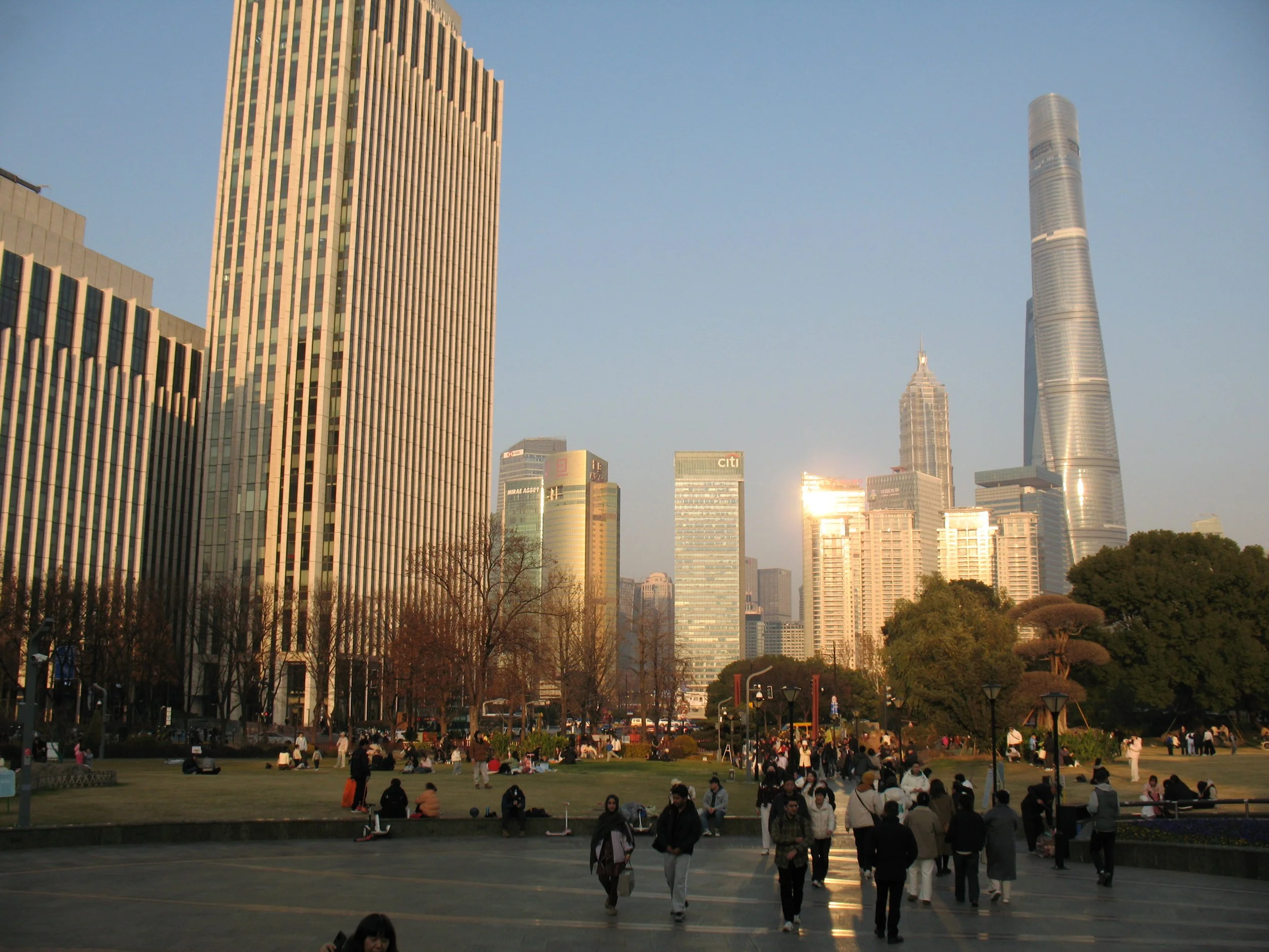 A cityscape with tall skyscrapers during sunset and people gathering in a park in the foreground.