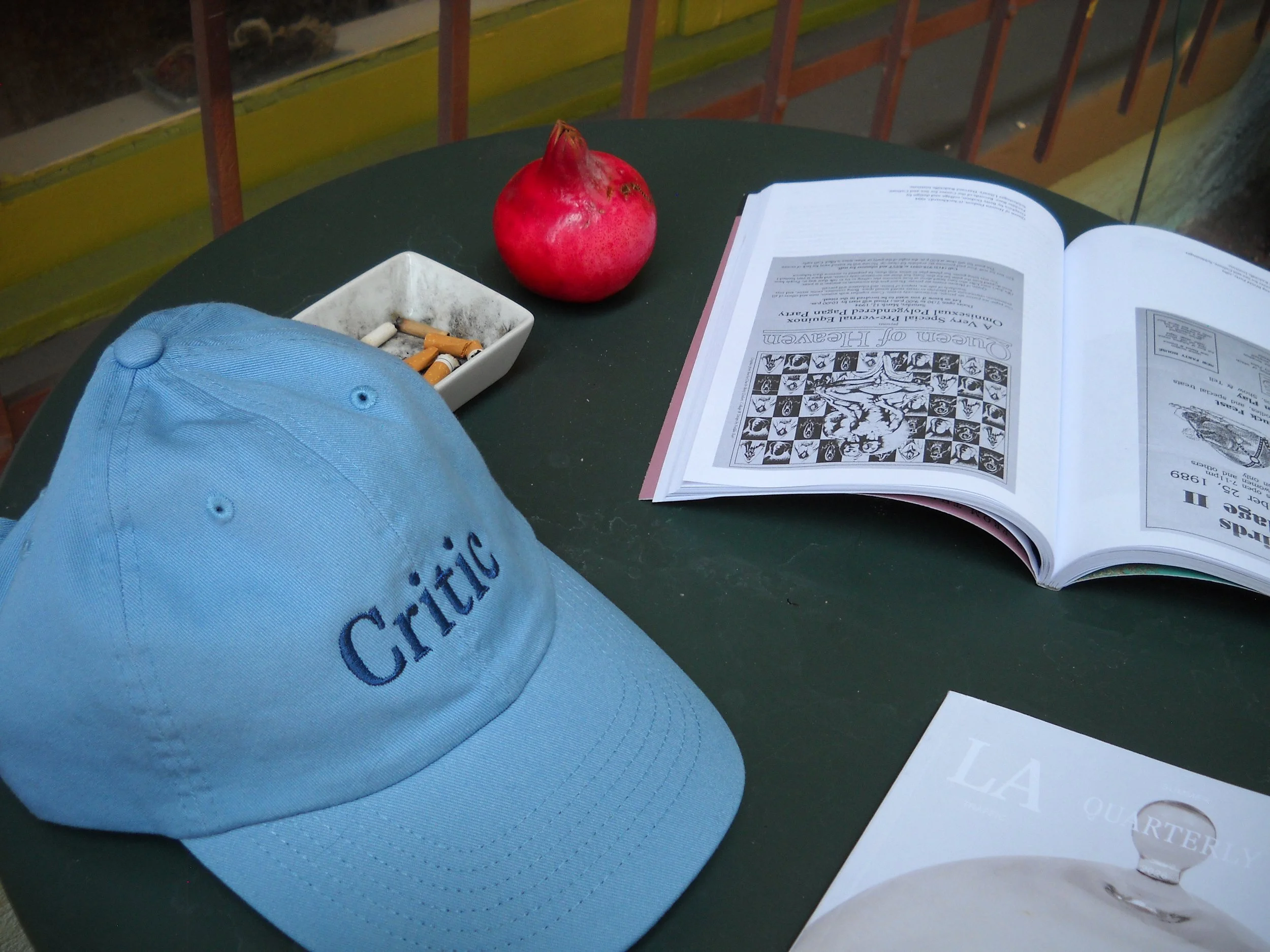 A blue baseball cap, a white ashtray with cigarette butts, an open book, a red pomegranate, and a paper with the text "Queens of Heaven" are placed on a green table.