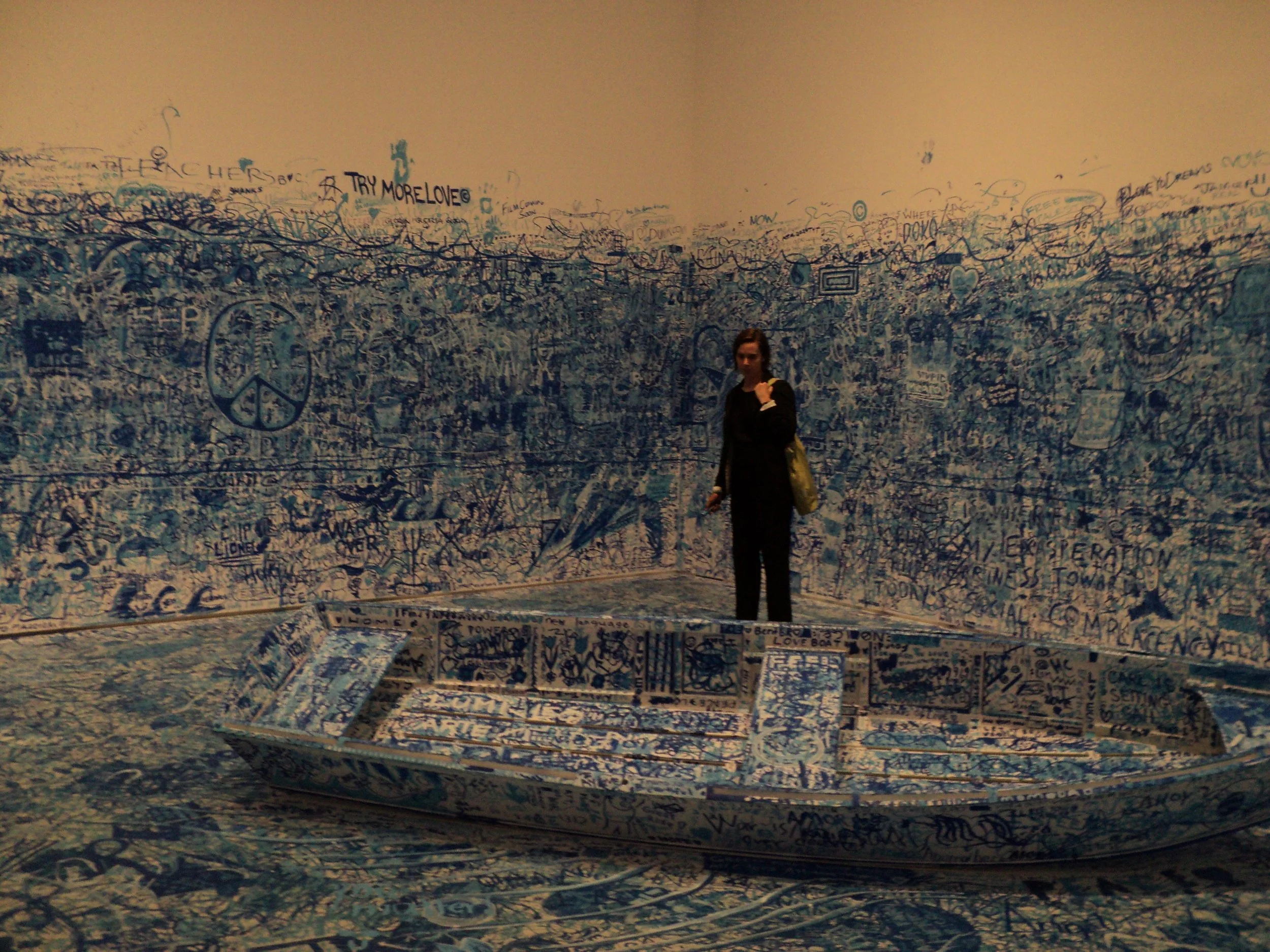 A woman standing in front of a large wall and floor covered entirely with graffiti and messages, some touching the ceiling, with a peace sign and the phrase 'TRY MORE LOVE' visible.