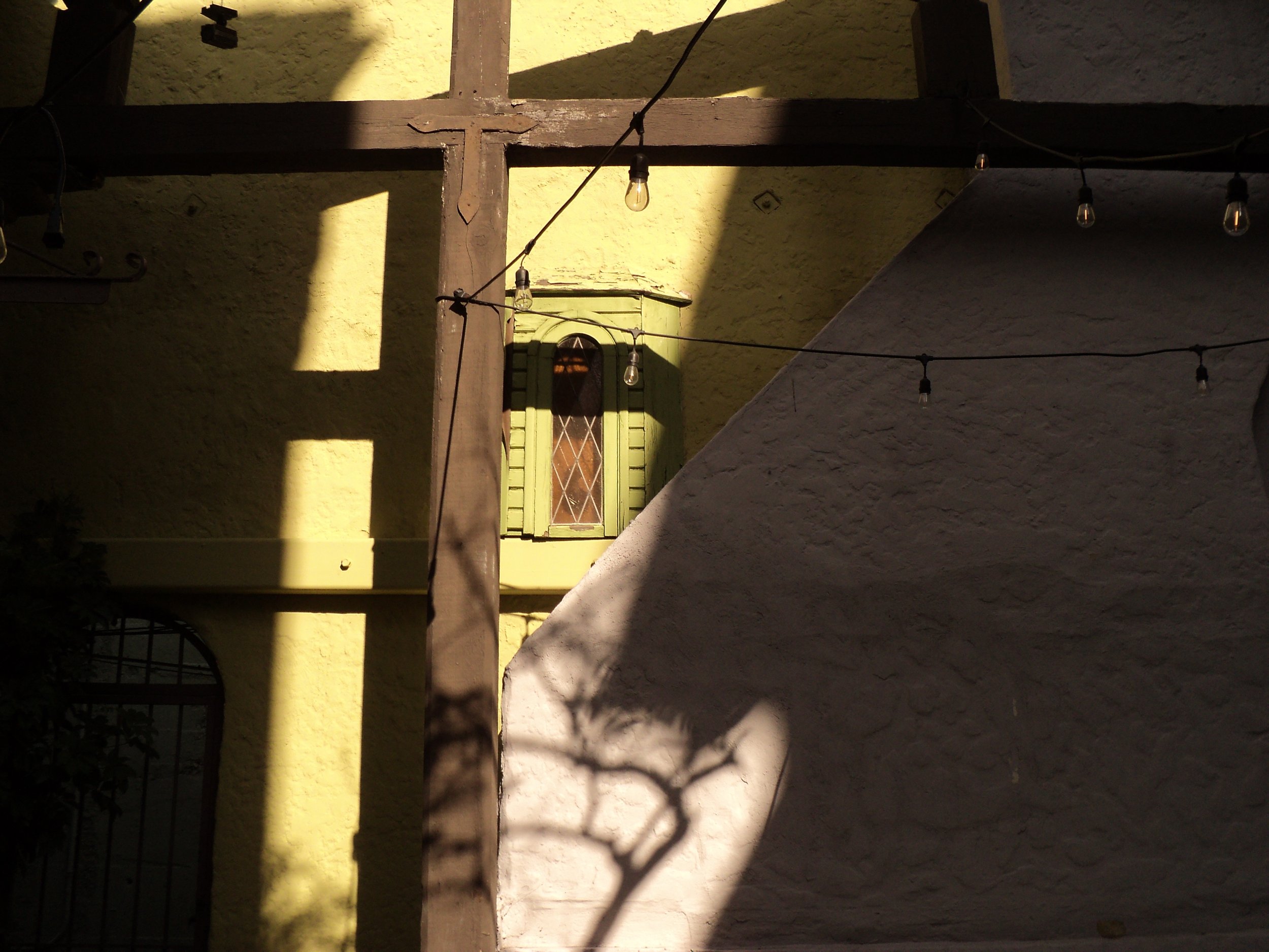 Close-up of a green window with shutter on a yellow wall, with string lights hanging in front and casting shadows.