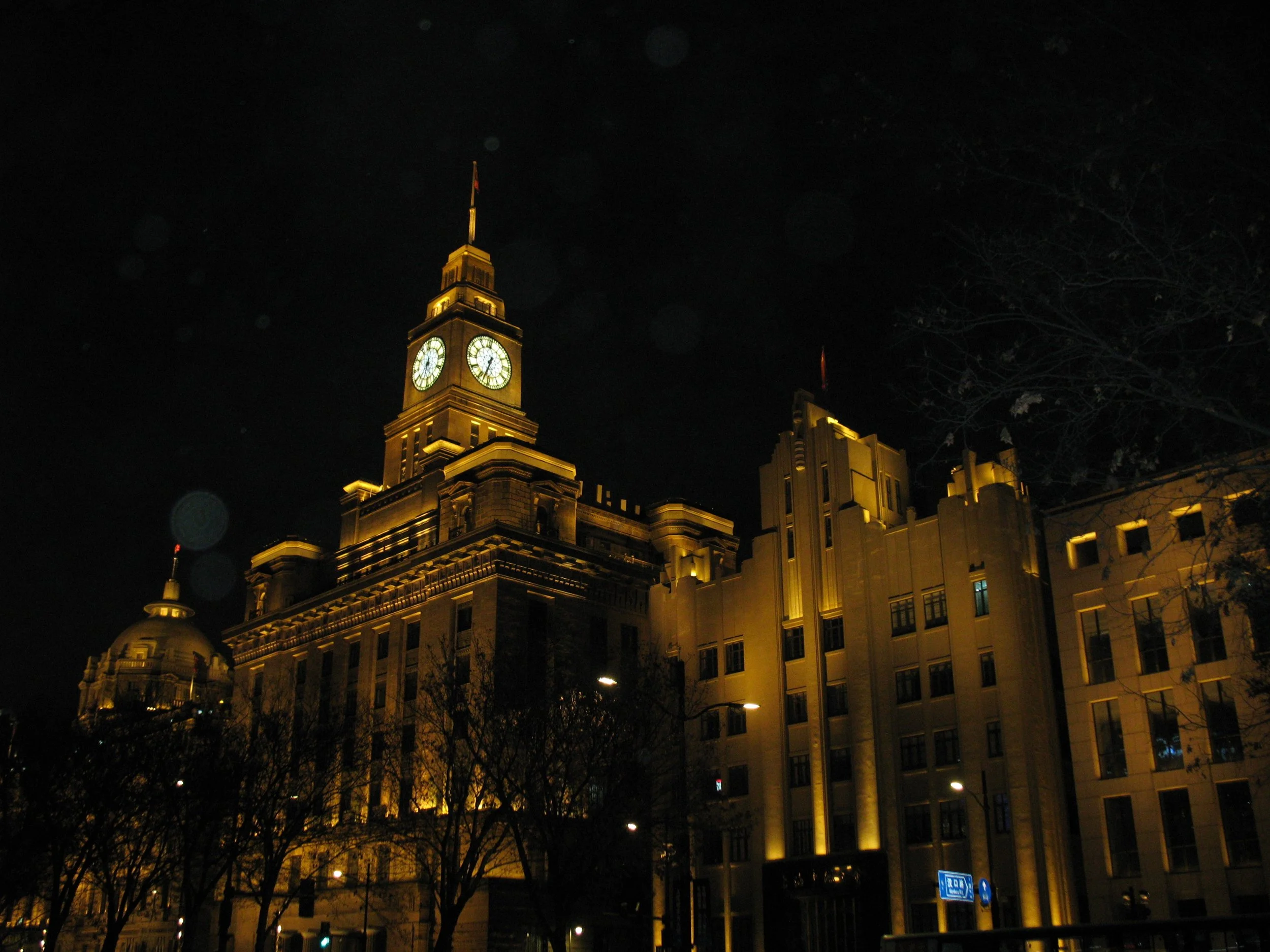 A lit historic clock tower building at night with a flag on top, surrounded by other buildings and leafless trees.