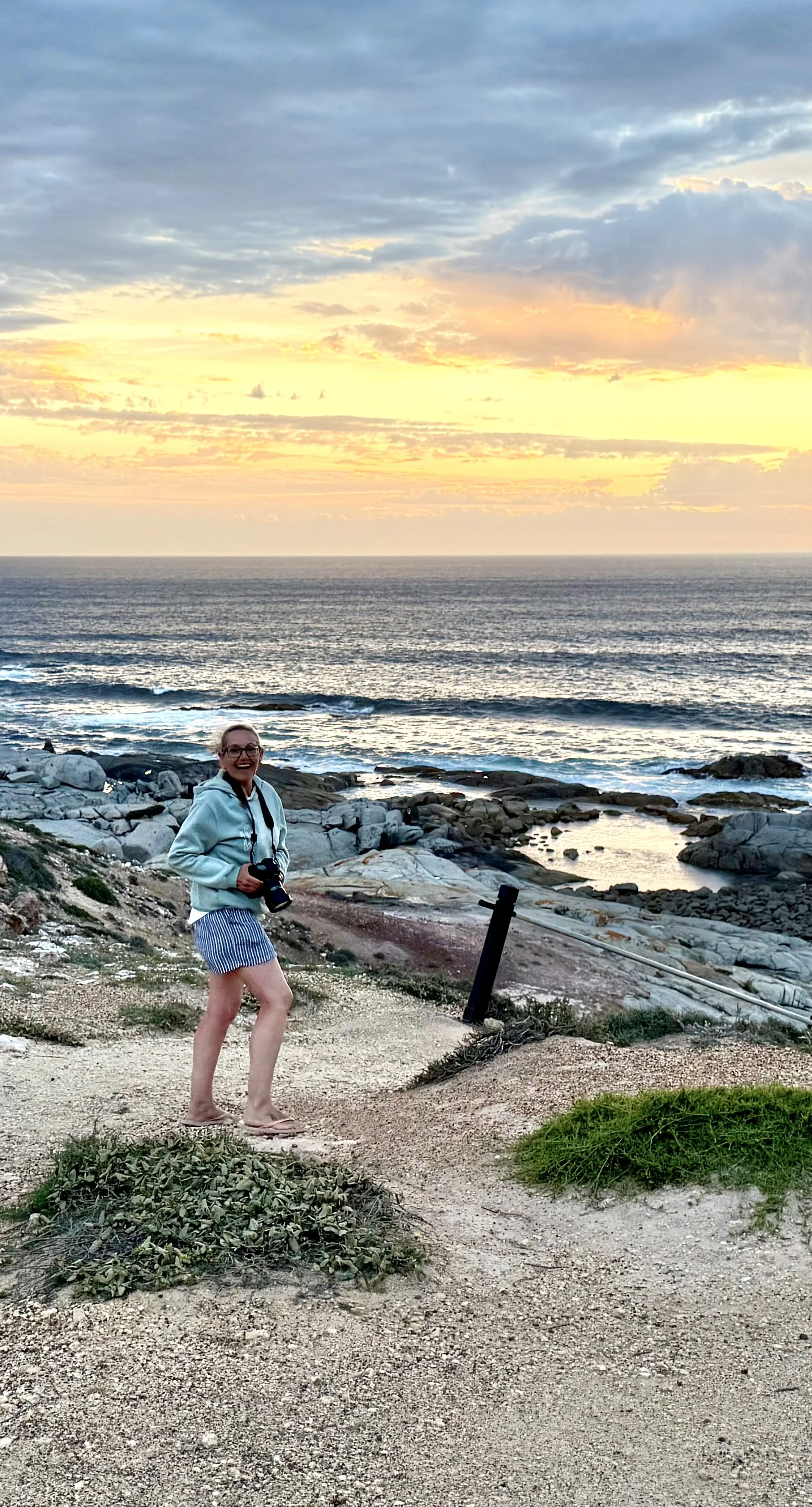A woman standing on a trail by the ocean during sunset, holding a camera, smiling, wearing glasses, a light blue hoodie, and striped shorts, with rocky shore and the sea in the background.