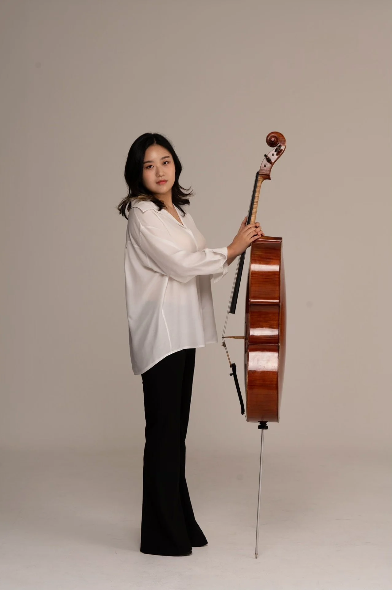 Young woman in white blouse and black pants holding a cello in a studio with neutral background.