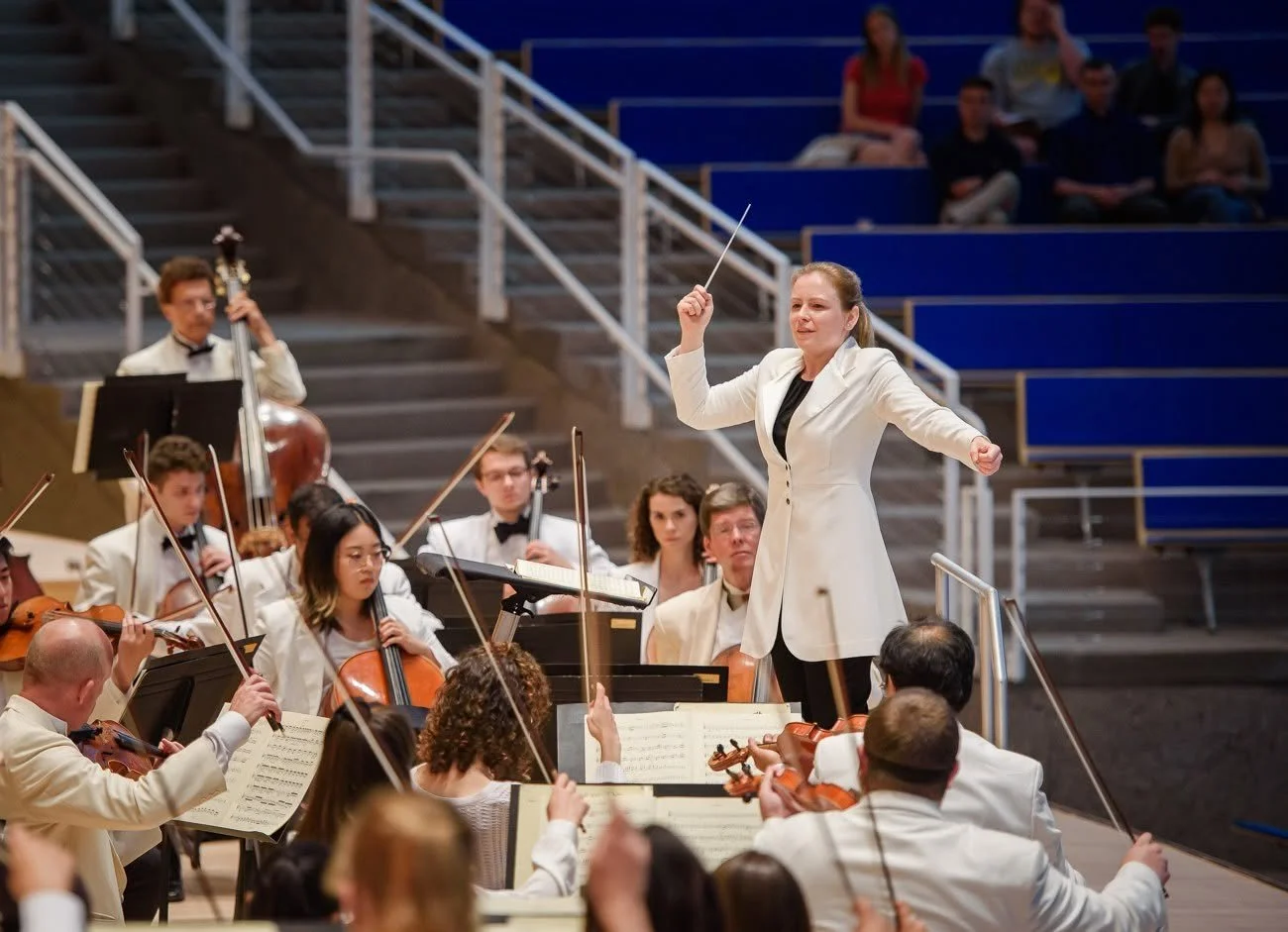 A woman conductor leading an orchestra during a performance, with musicians playing strings and an audience in the background.