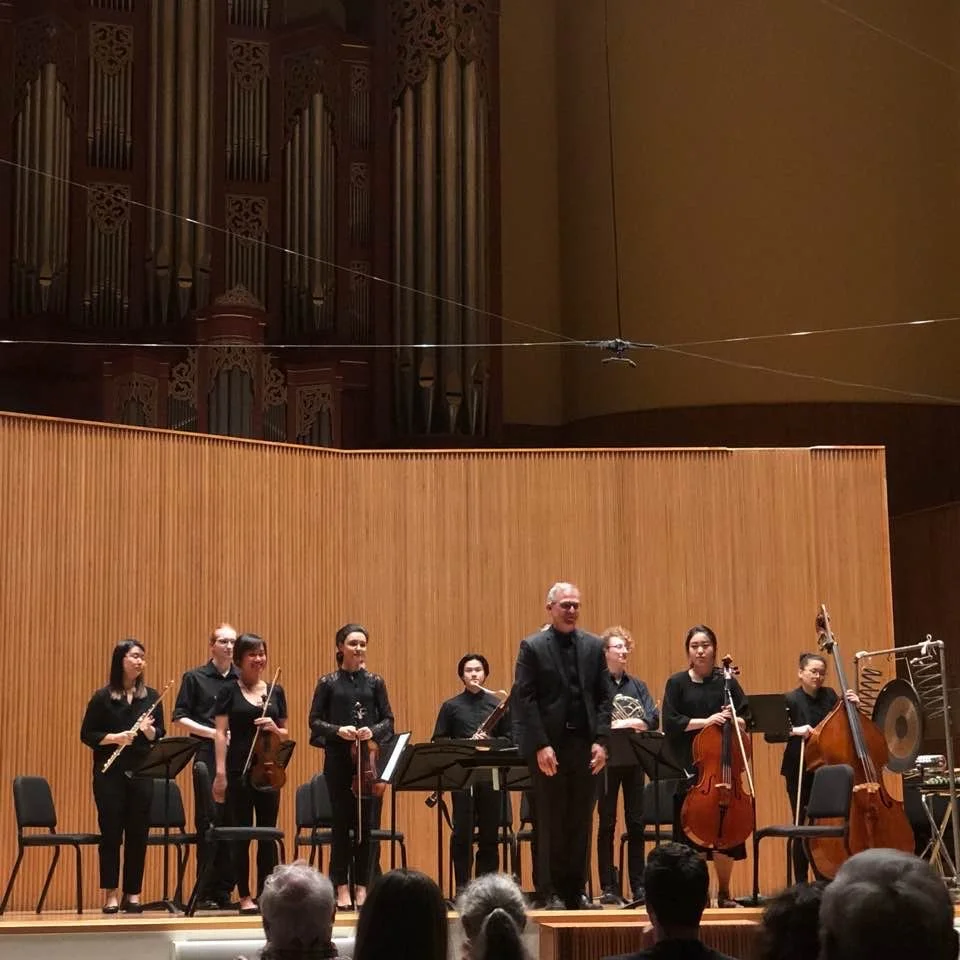 Orchestra on stage after a performance, with seven musicians holding instruments including violin, viola, cello, bass, flute, and a gong, and a conductor standing in front of them, in a concert hall with wood paneling and pipes of an organ in the bac