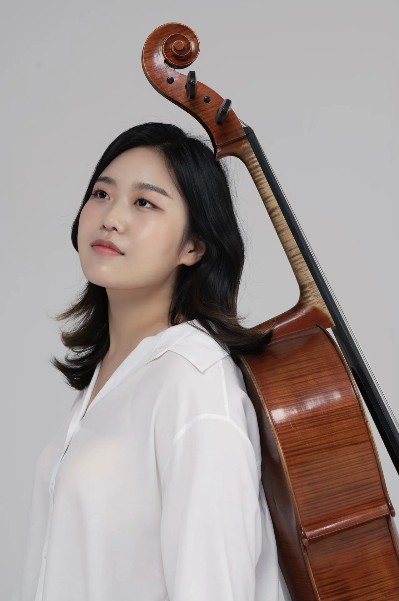 A woman with shoulder-length black hair and light skin, wearing a white shirt, is balancing a wooden cello on her shoulder against a plain gray background.