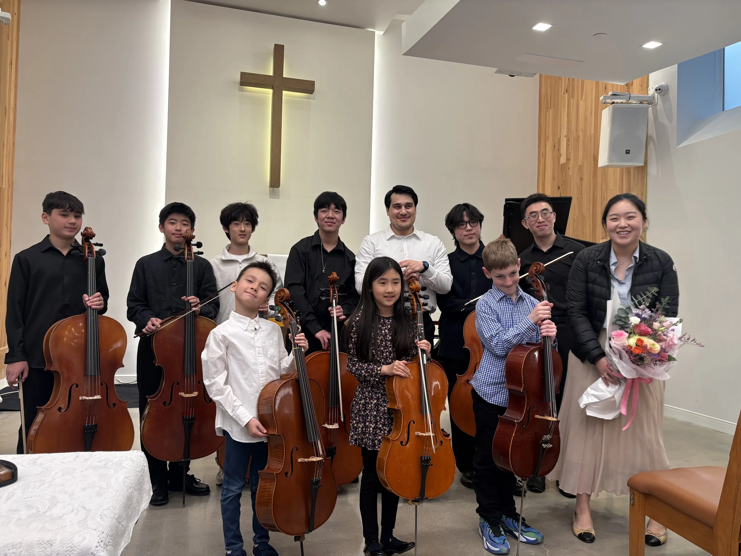 Group of young musicians holding cellos and posing for a photo in a church with a cross on the wall behind them.