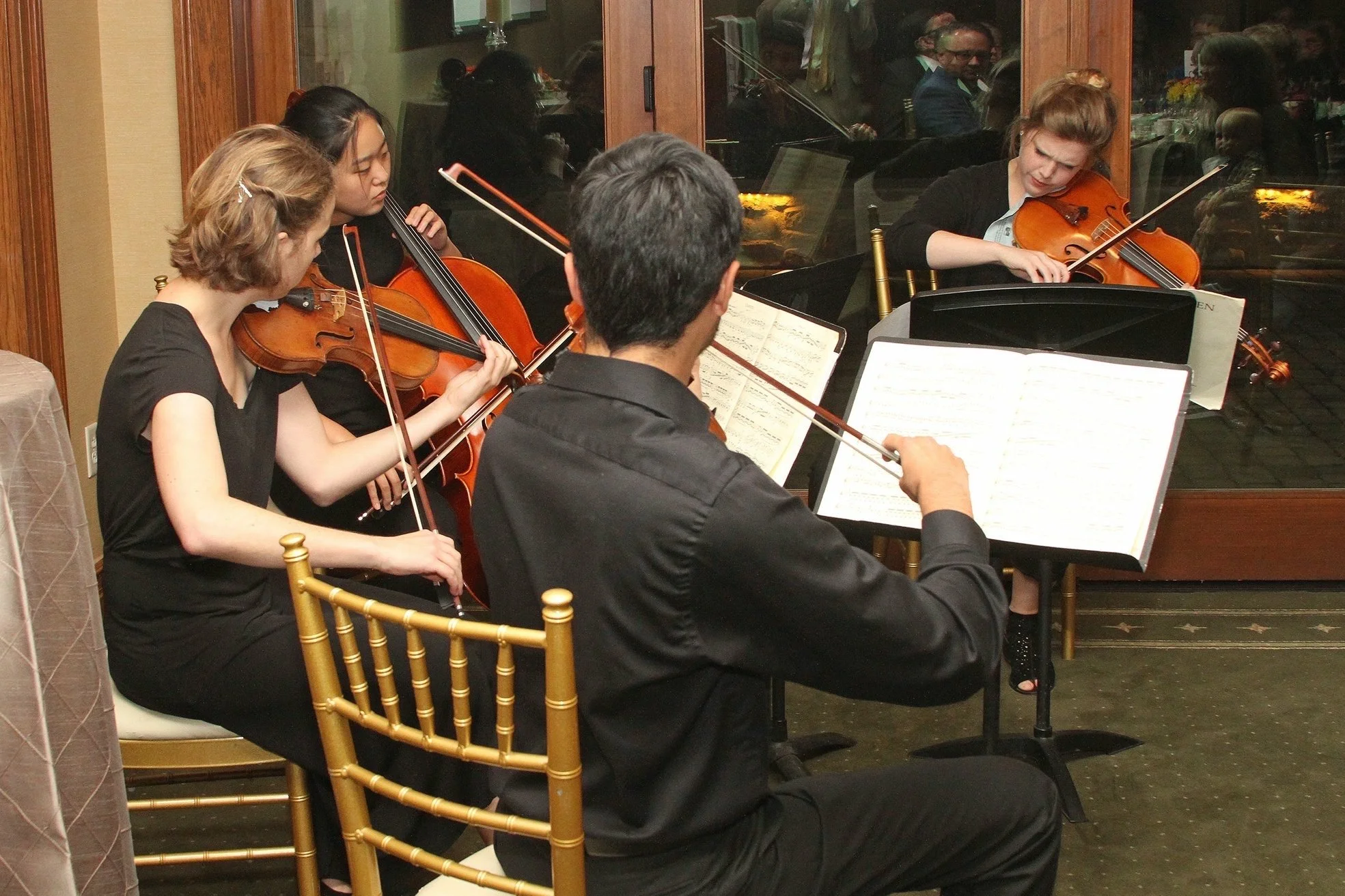 A small string quartet performs in a restaurant, seen through a glass window. The group includes two women on violins, a woman on a cello, and a man on a violin, all reading sheet music while playing. Reflection of spectators and piano can be visible