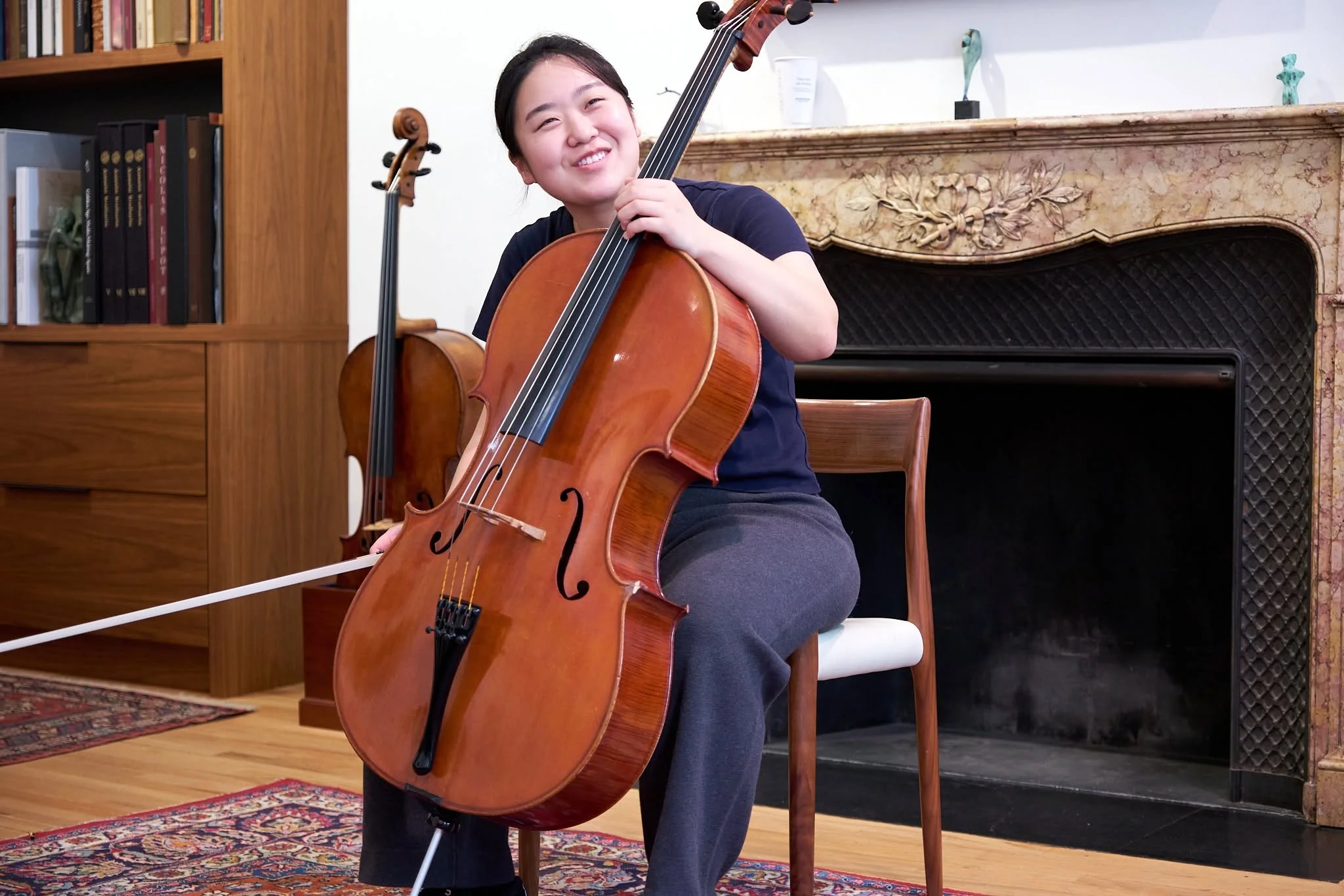 A young woman smiling while playing a cello in a room with wooden furniture and bookshelves.