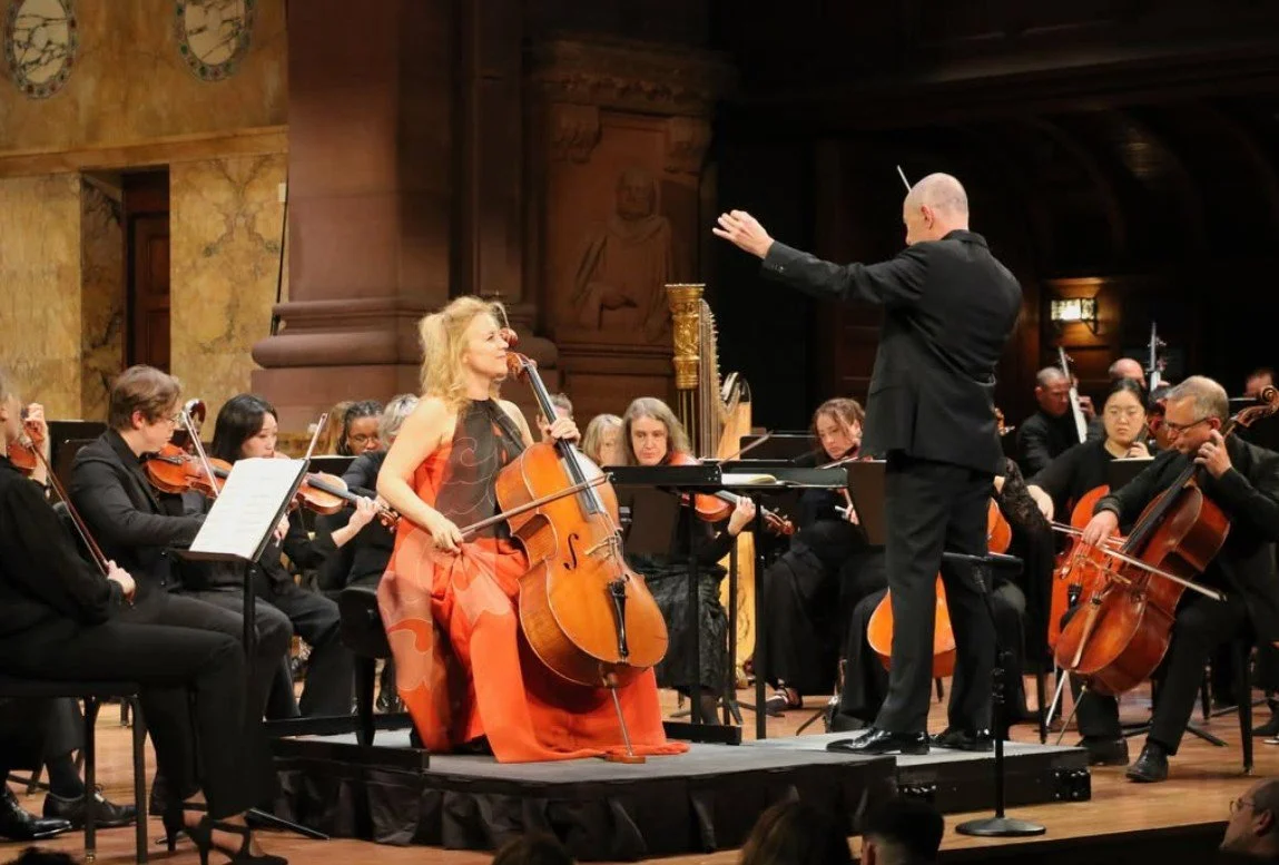 Orchestra performing in a concert hall with a female cellist in an orange dress and a male conductor leading the group.