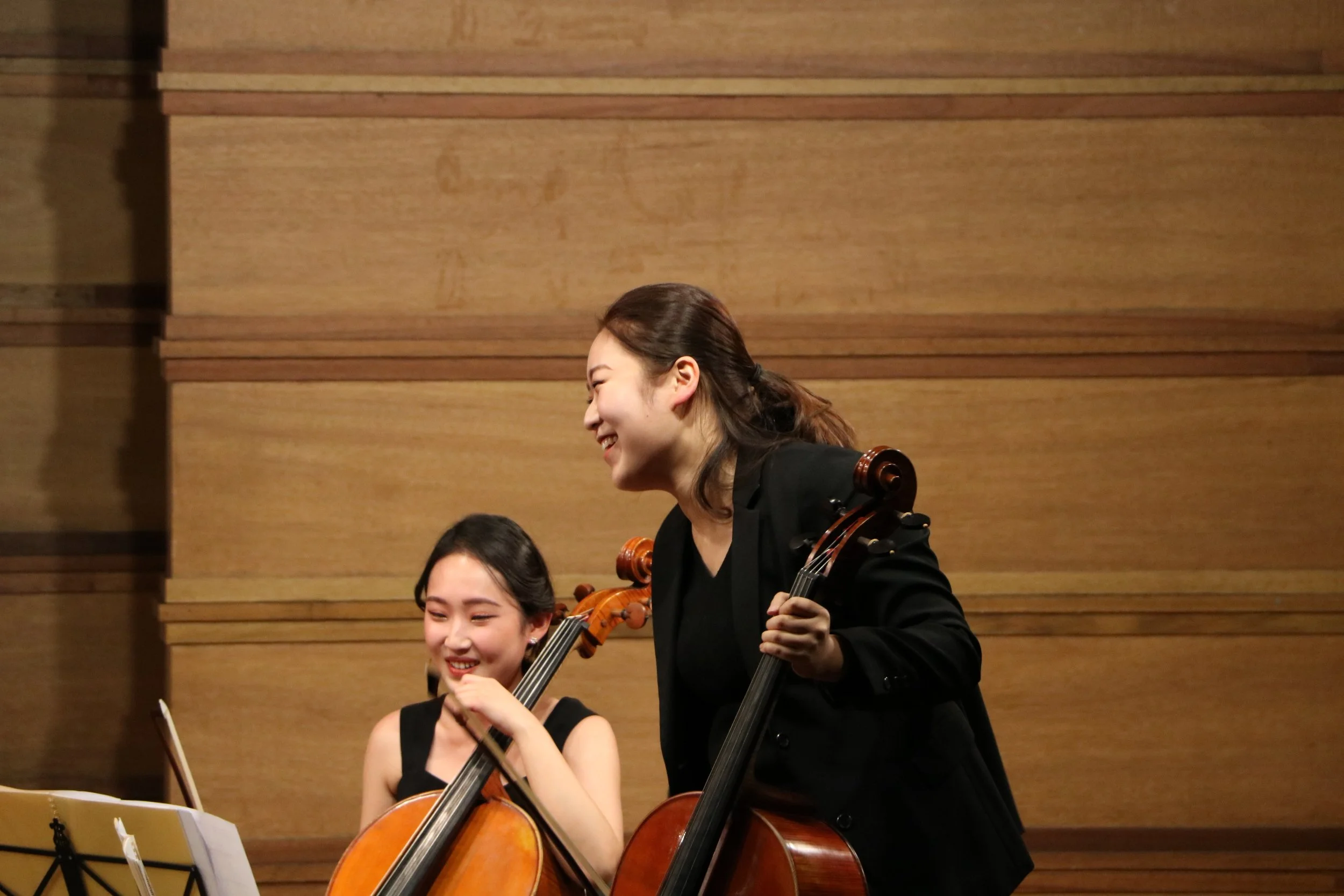 Two women playing cellos in a performance hall, smiling and enjoying their music, with a wooden wall background.