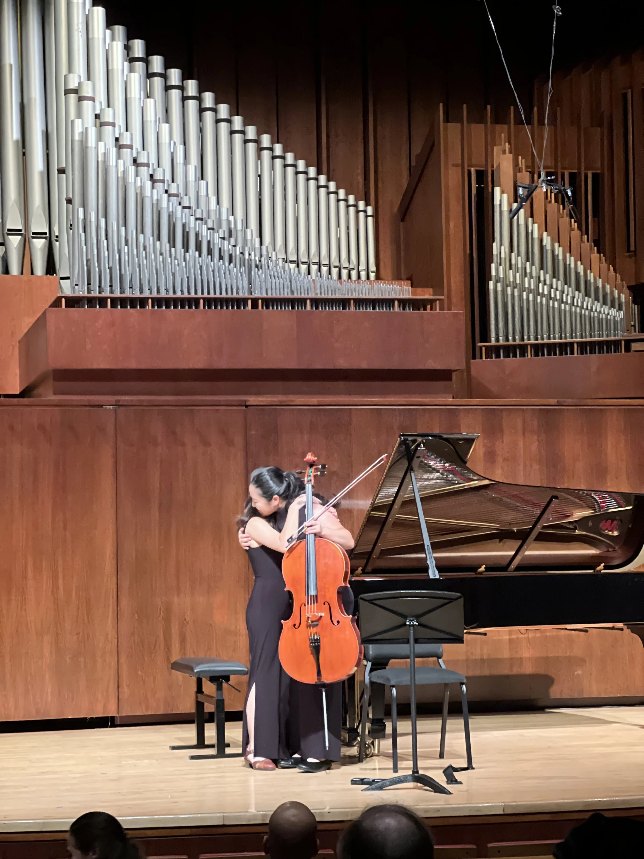 A woman is hugging a cello on stage in front of a grand piano, with large organ pipes in the background.