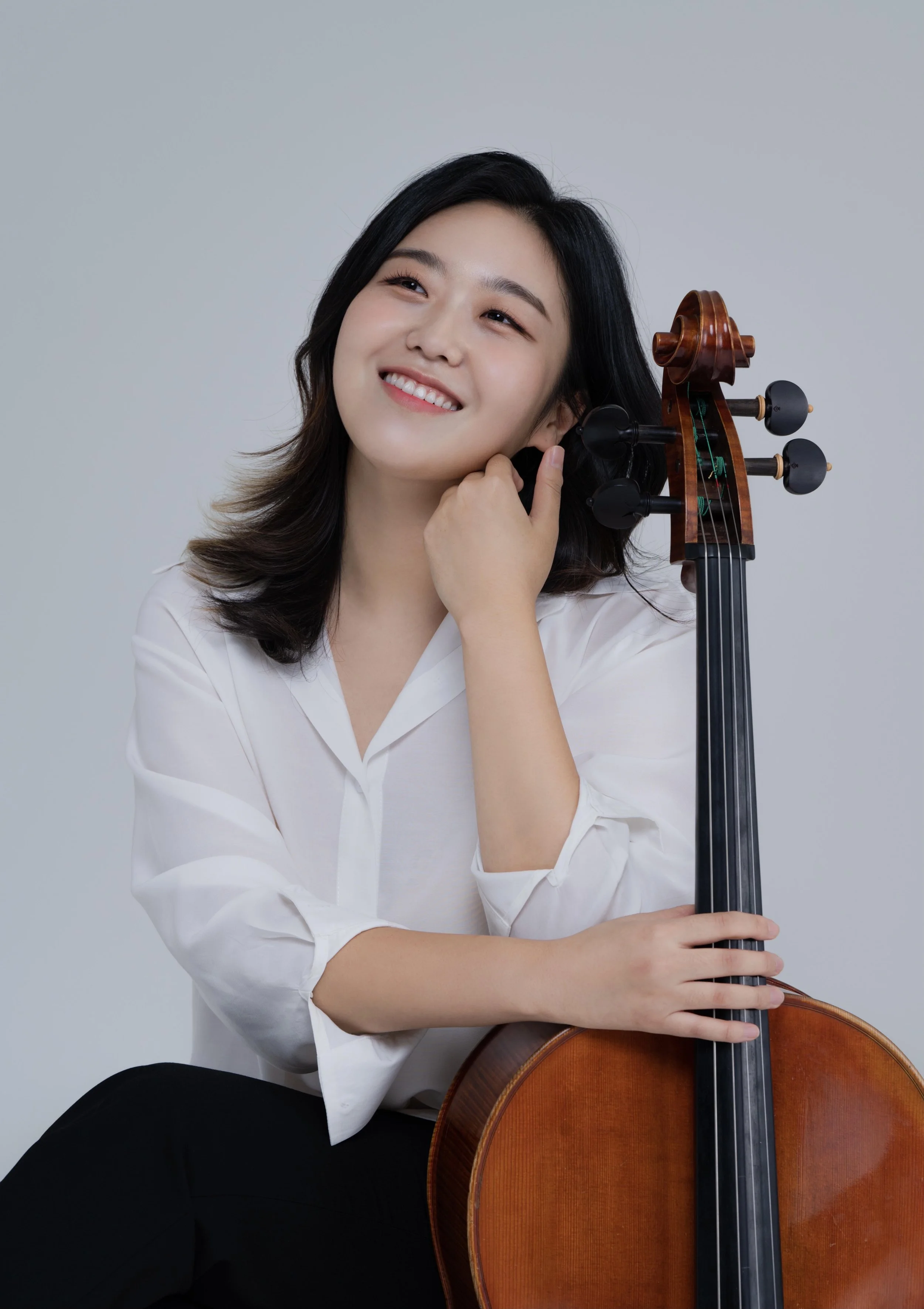 A young woman with dark hair smiling and sitting with a cello, wearing a white blouse and black pants, against a plain gray background.
