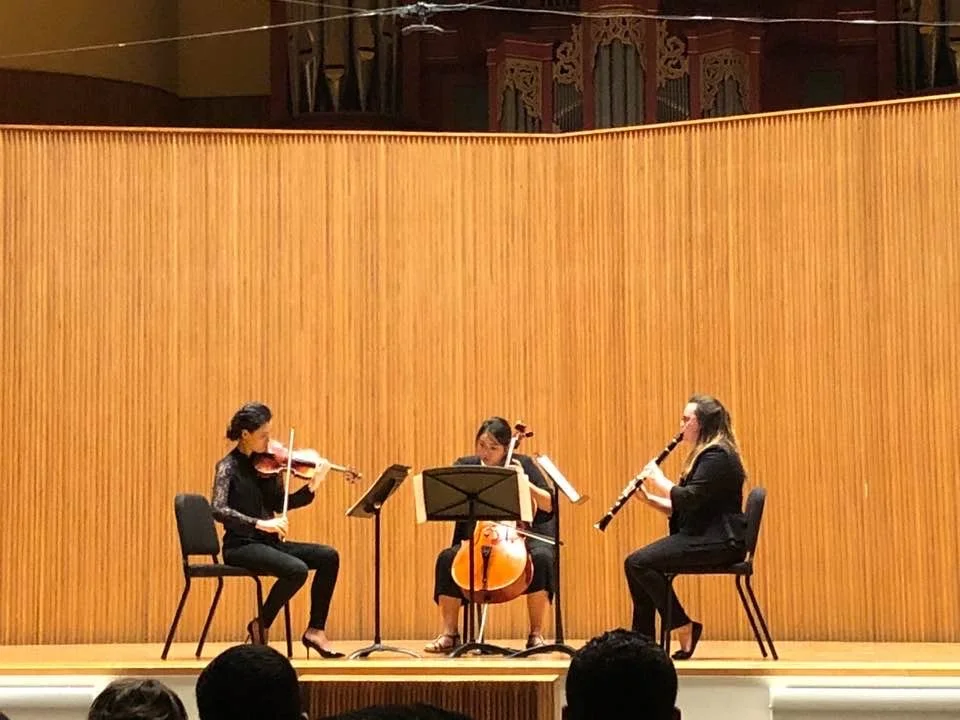 Three women are performing a classical music piece with a violin, cello, and clarinet on stage in a concert hall with a wooden wall background.