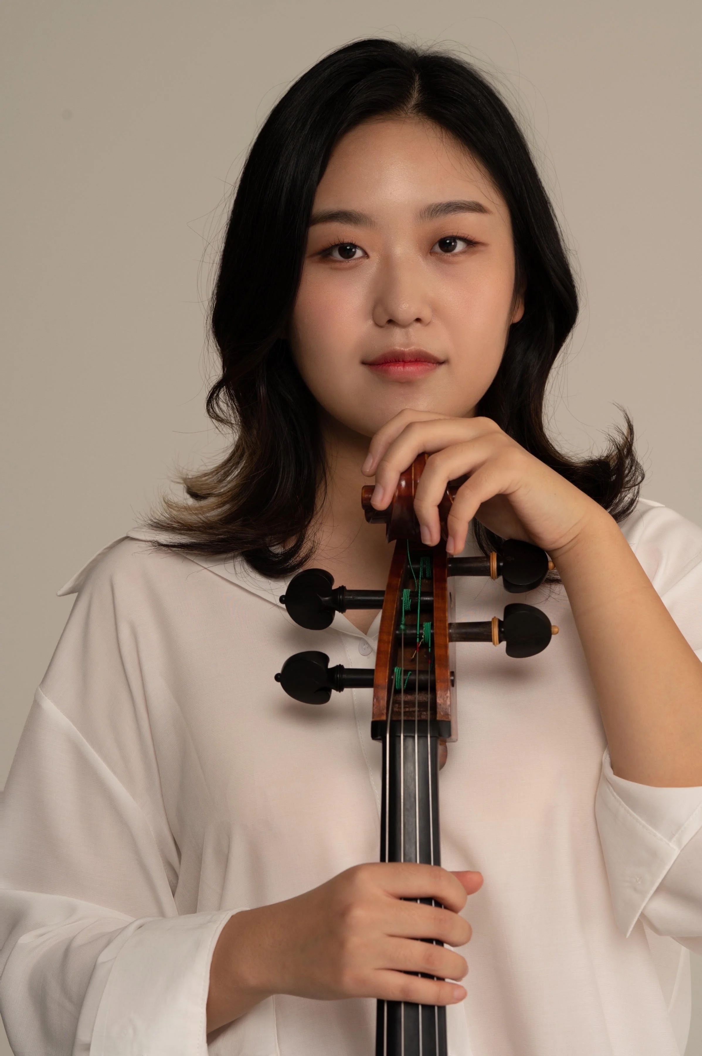 A young woman with black hair and a white shirt holding a violin indoors against a neutral background.