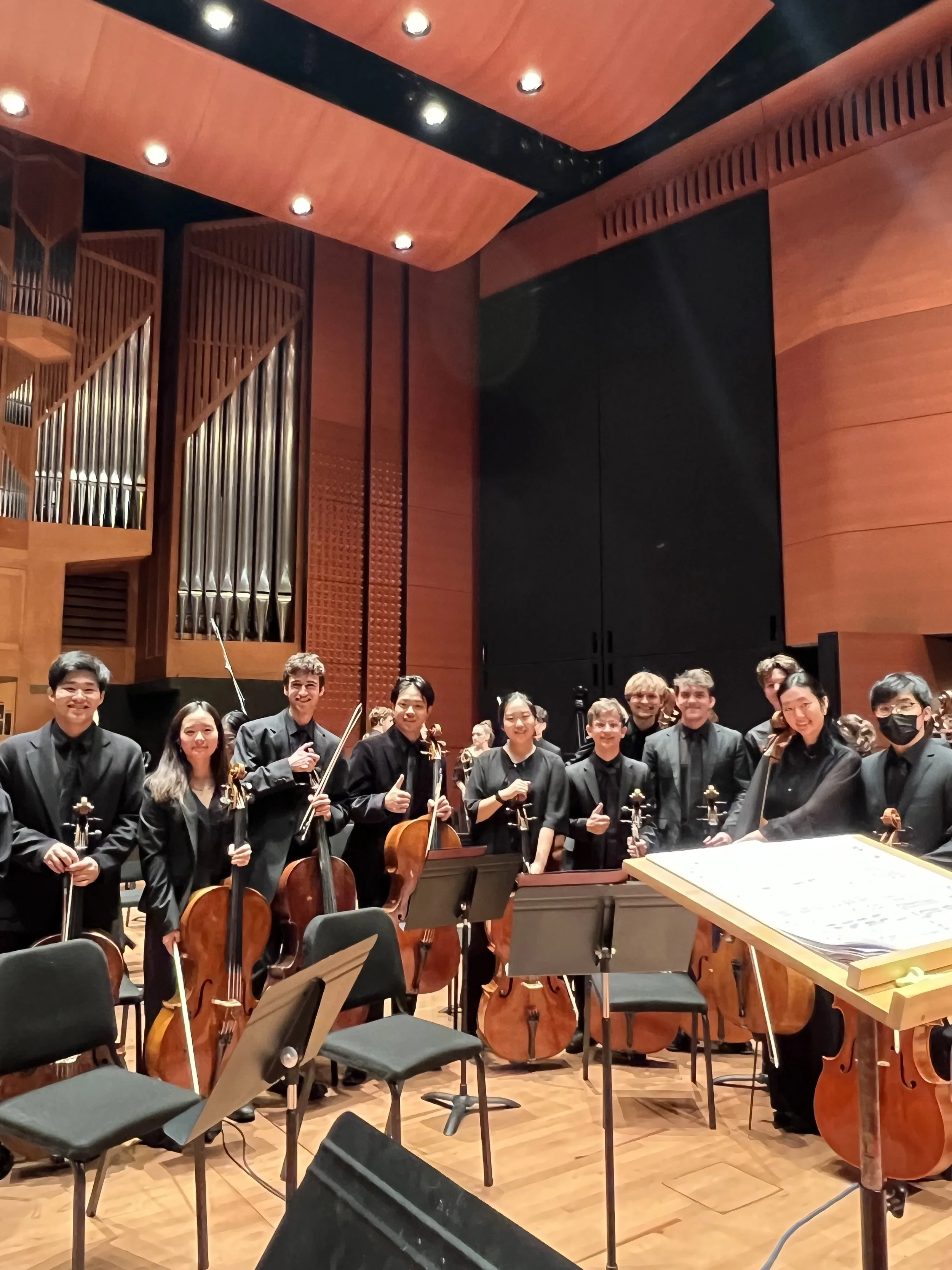 A group of young musicians in formal black attire holding string instruments such as violins and cellos, standing in a concert hall with wooden walls and musical equipment, smiling and posing for the photo after a performance.