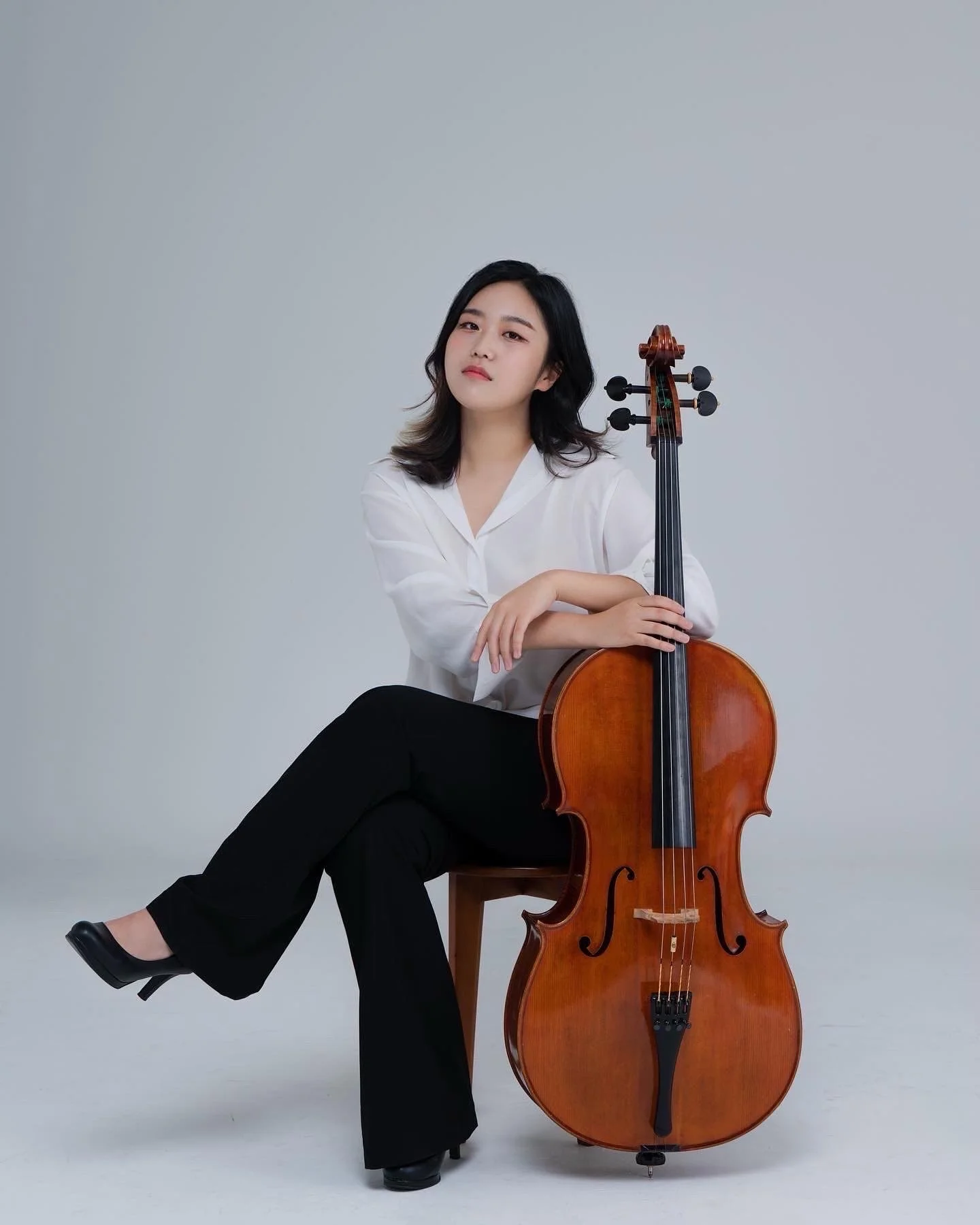A young woman with black hair and a white blouse sitting cross-legged on a wooden stool, holding a cello upright, against a plain gray background.