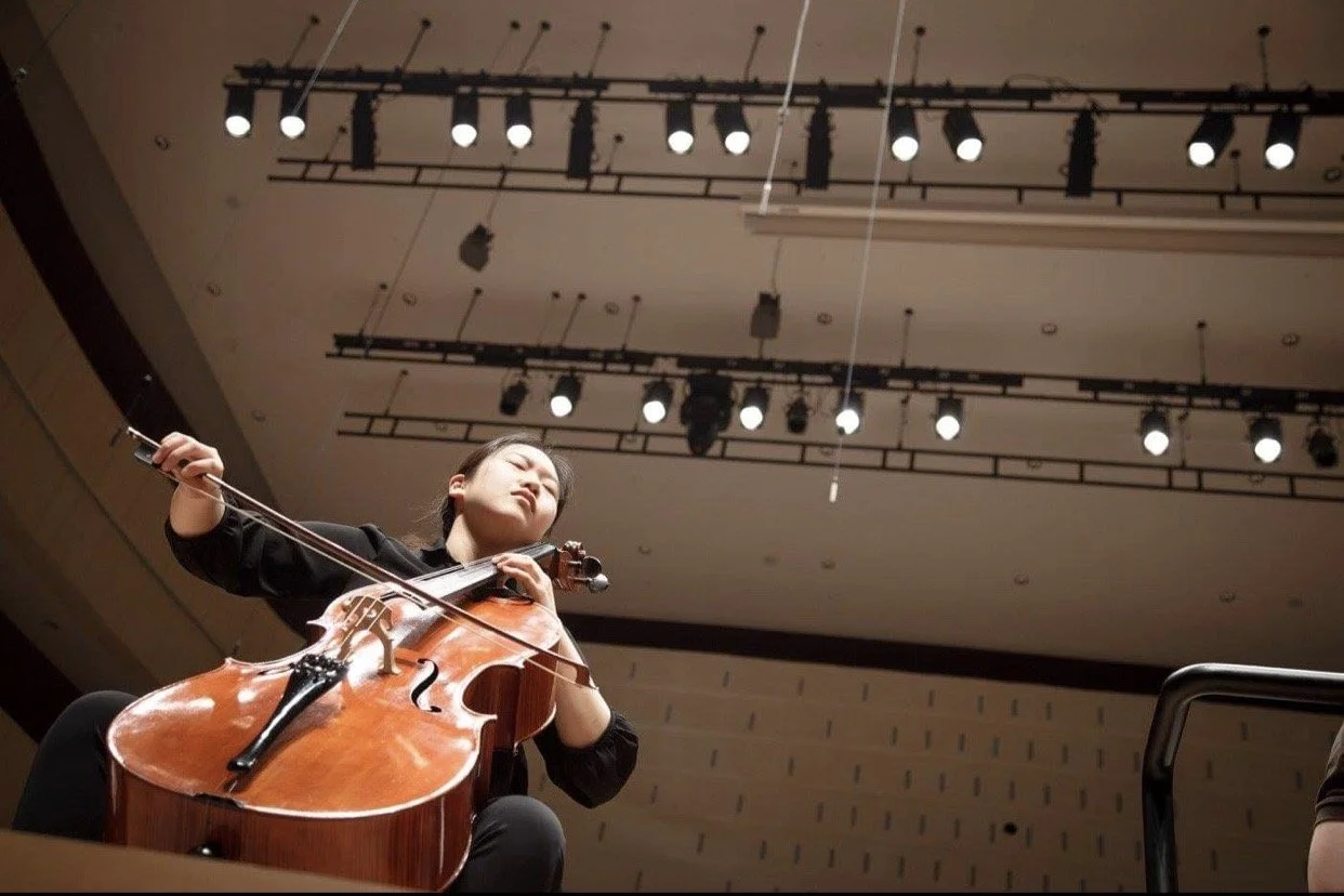 A woman playing a cello on a stage in a concert hall with stage lights overhead.