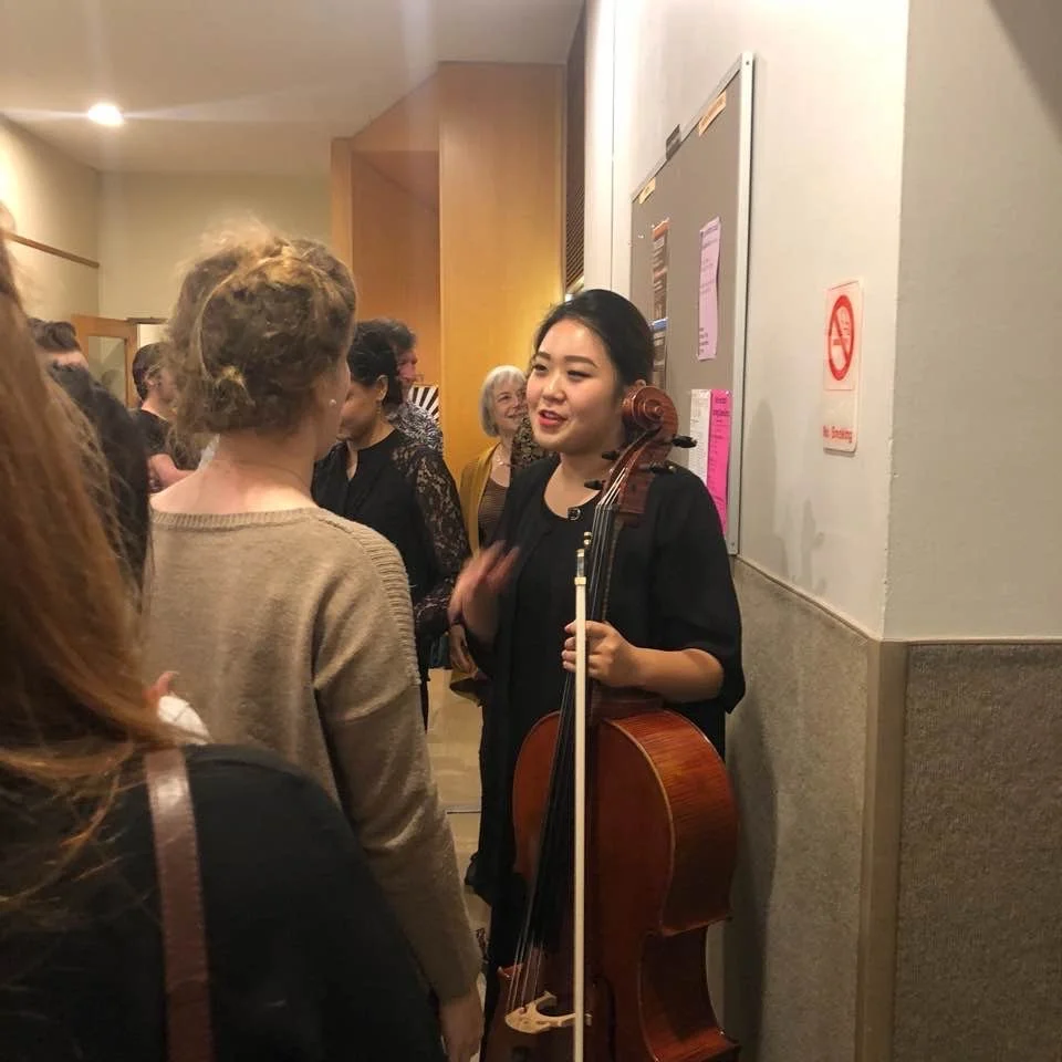 A woman with dark hair holding a cello and bow, talking to a group of women in a hallway or lobby.