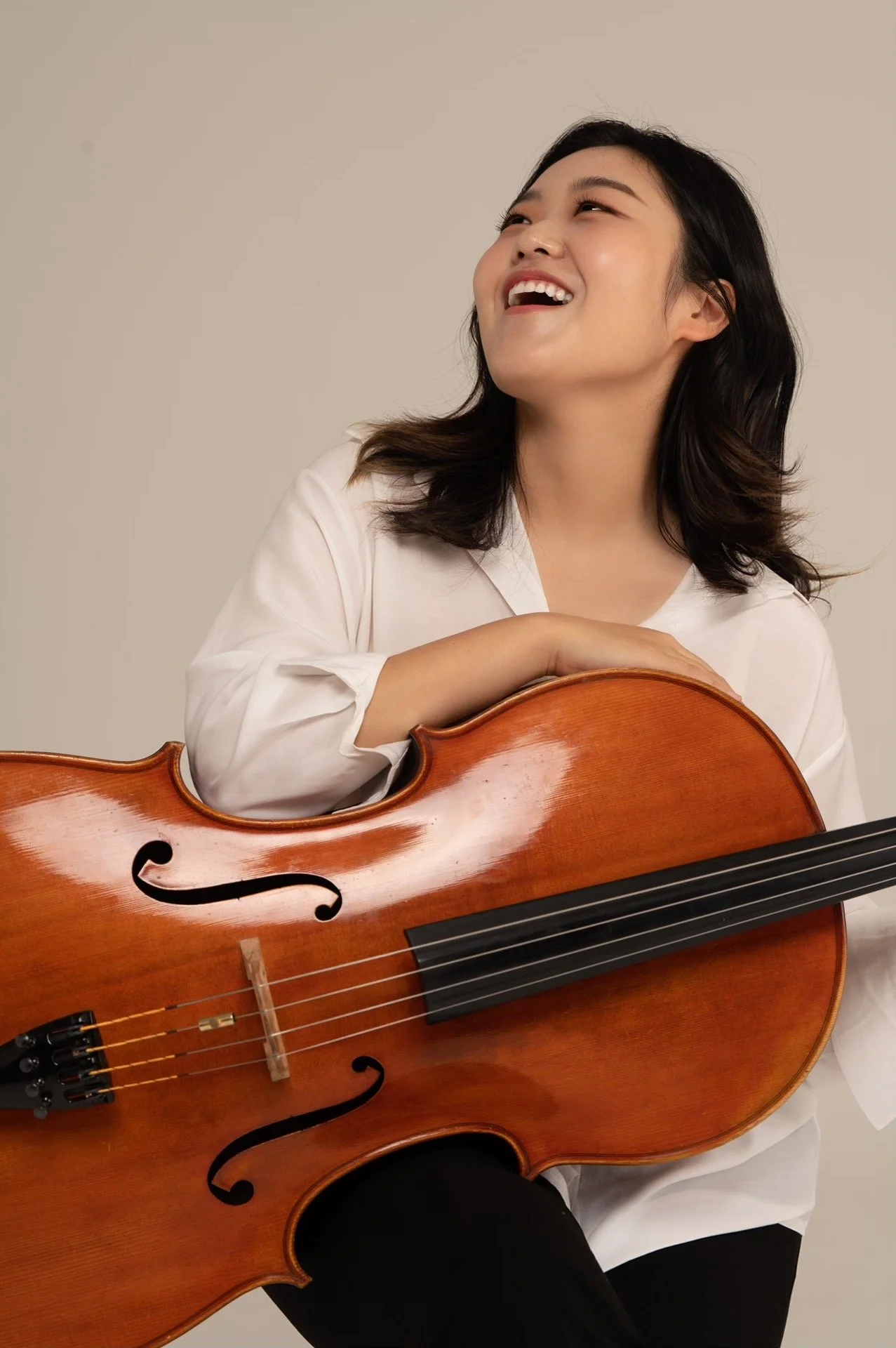 A woman with shoulder-length dark hair smiling and looking up, sitting behind a wooden cello, wearing a white shirt, against a plain beige background.