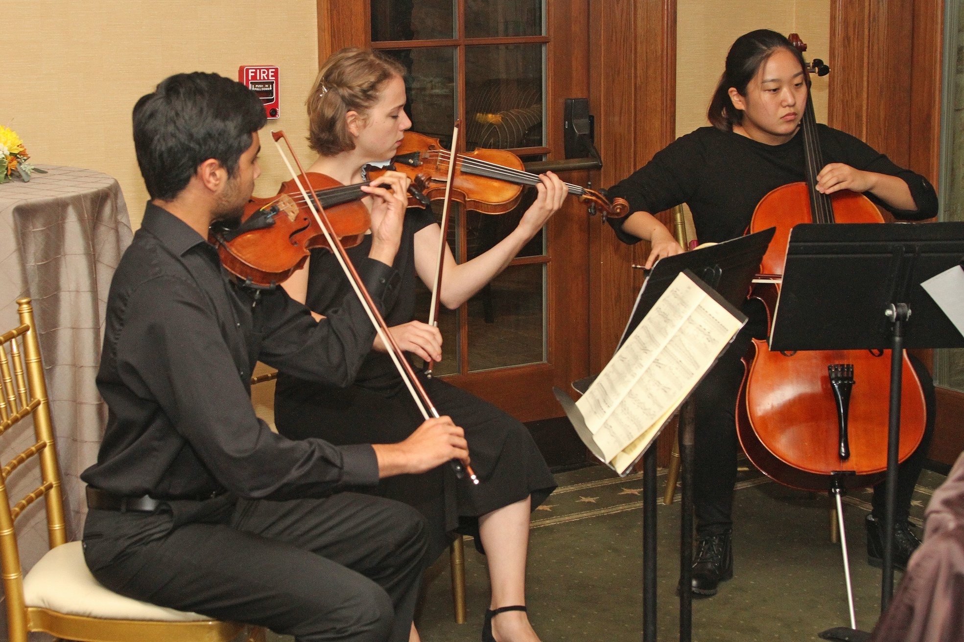 Three musicians playing string instruments at an indoor event: a man playing a violin, a woman playing a violin, and another woman playing a cello, with sheet music on stands in front of them.