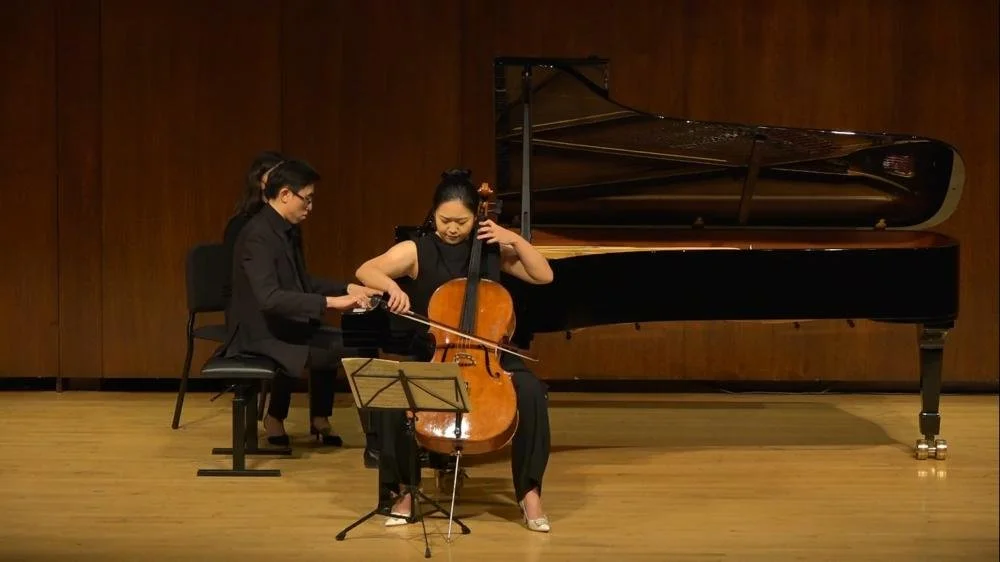 Two women playing piano and cello on stage with a grand piano in the background.