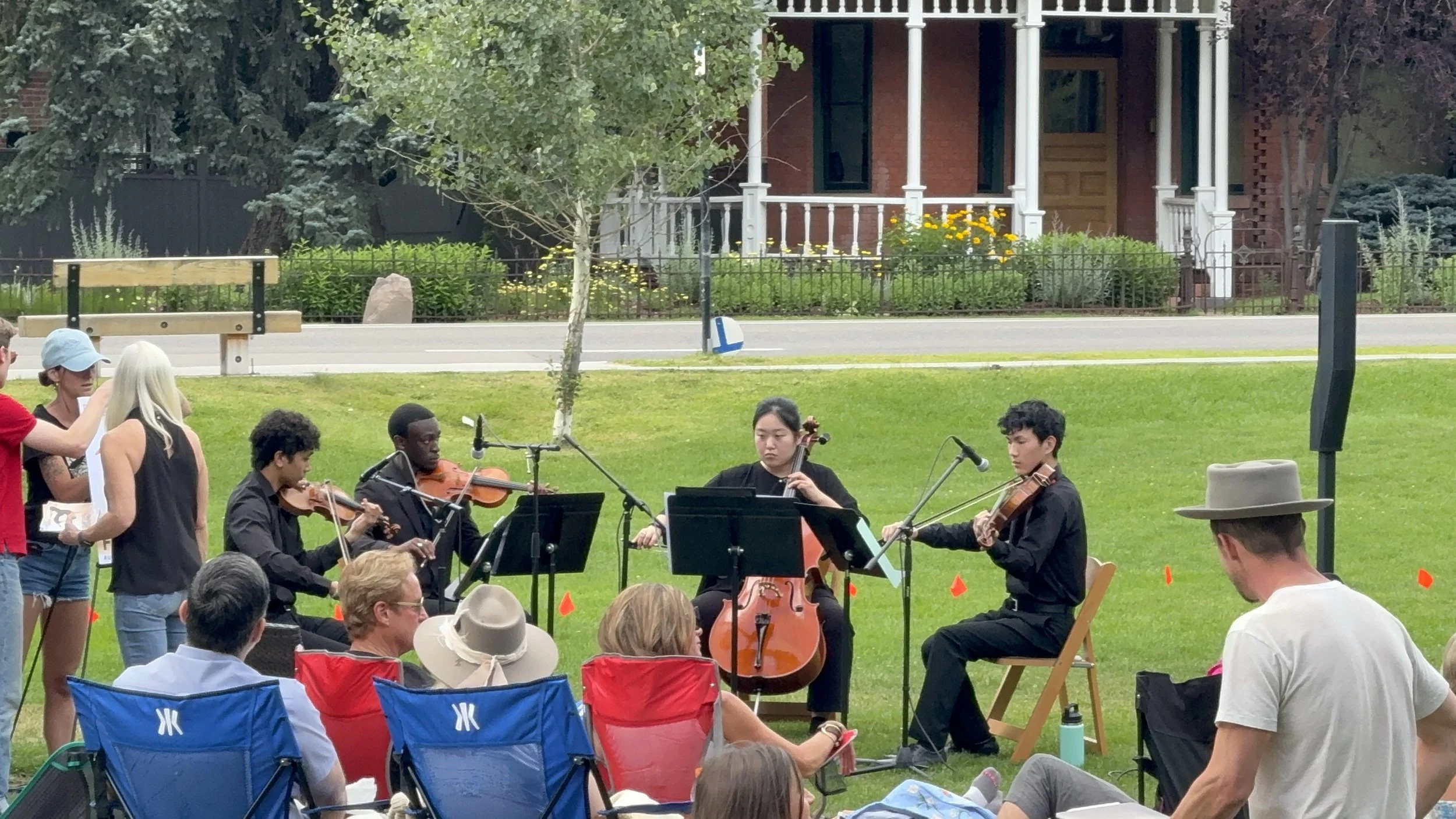 A group of young musicians playing string instruments including violins and a cello at an outdoor concert, with an audience seated on folding chairs on a grassy field. The scene includes a person conducting, some standing around, and a background of 