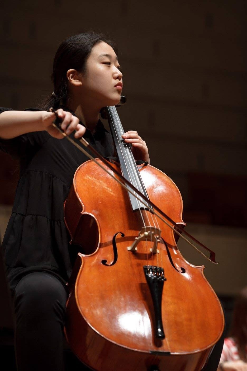 Young woman playing a cello, dressed in black, with focused expression in a dimly lit room.