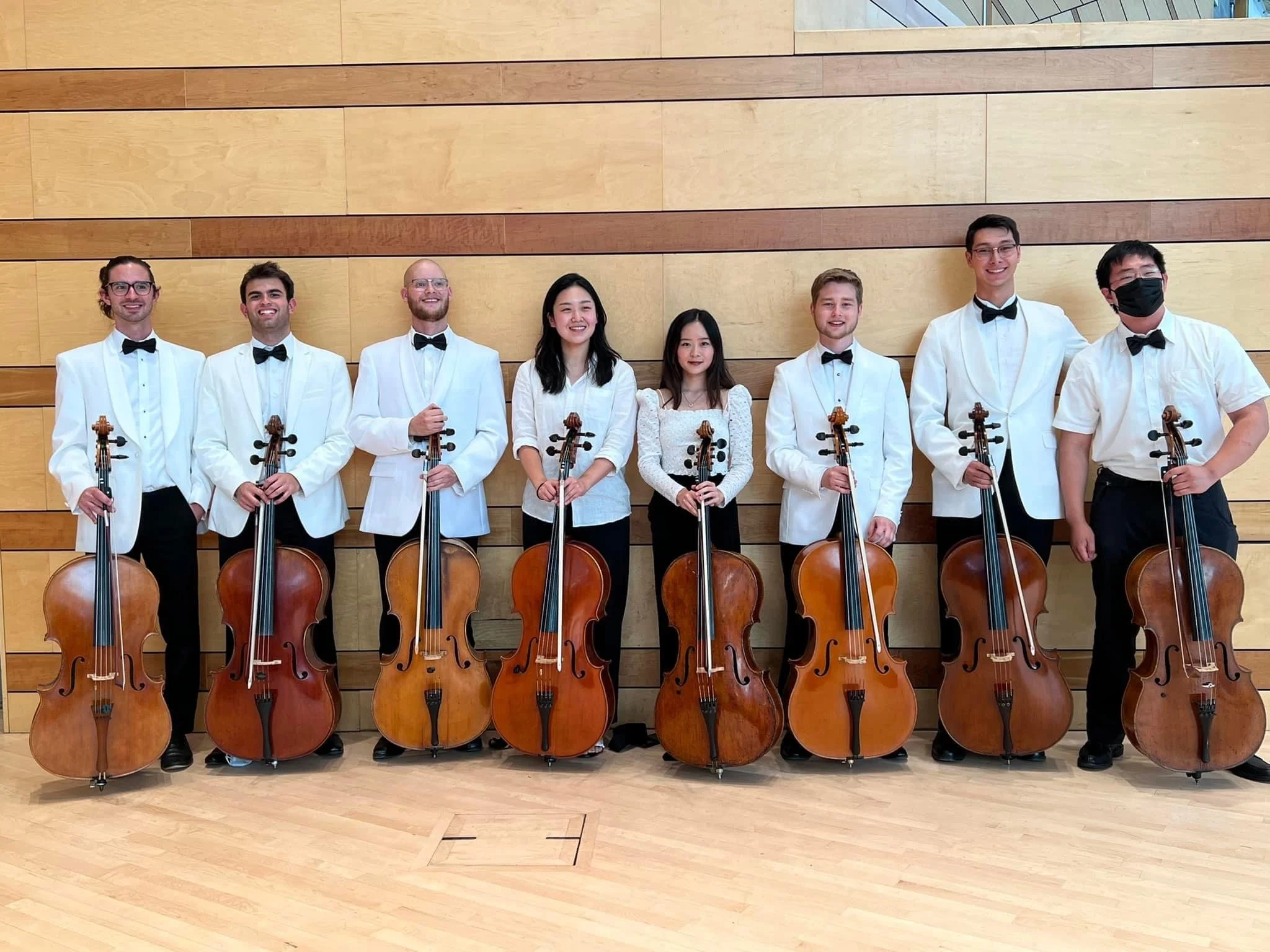 A group of eight musicians with violins standing in front of a wooden wall.