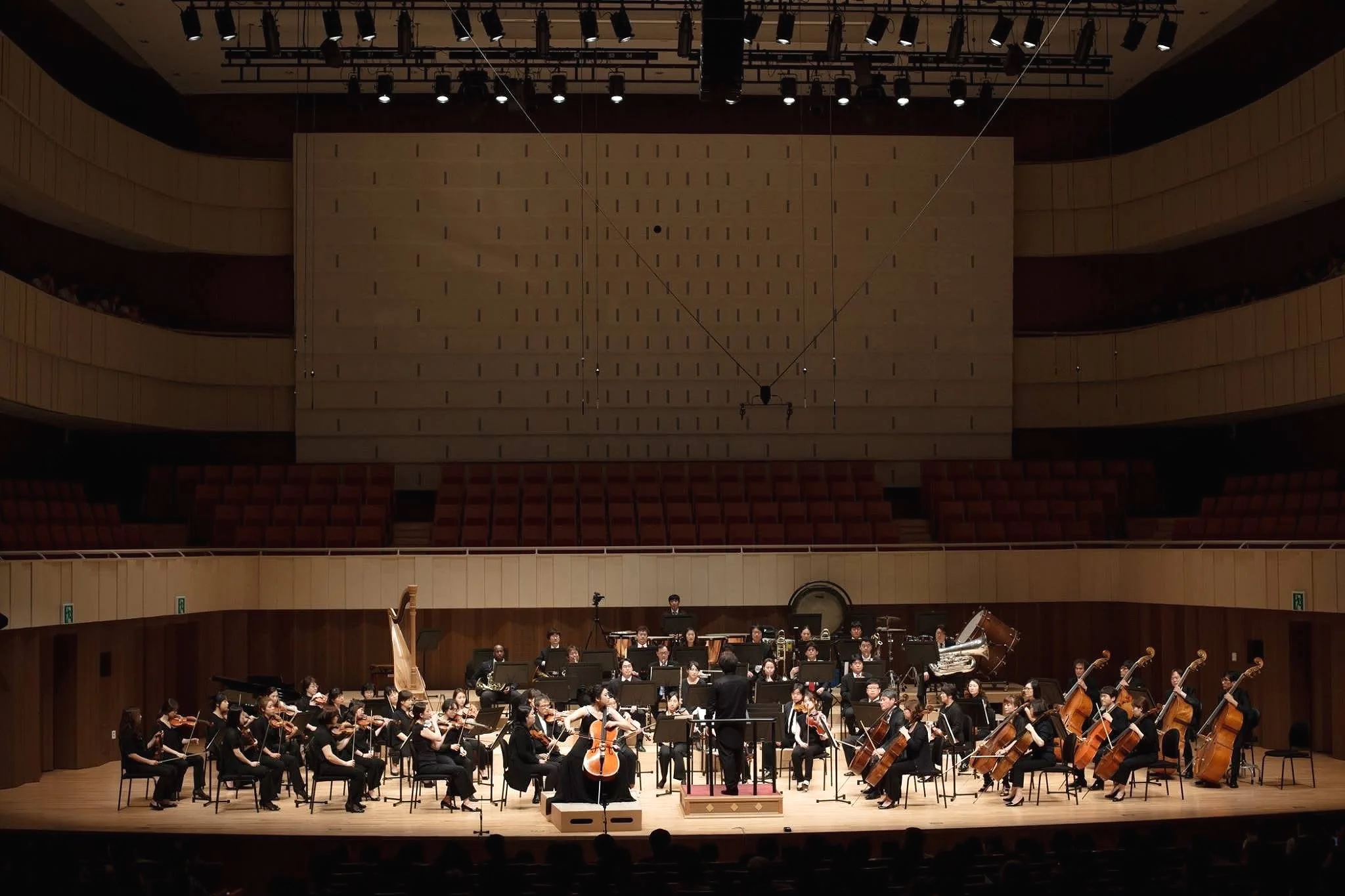 An orchestra performing on stage in a large concert hall with a wooden backdrop and balcony seating. Musicians are playing various instruments including strings, brass, and percussion, under stage lighting.