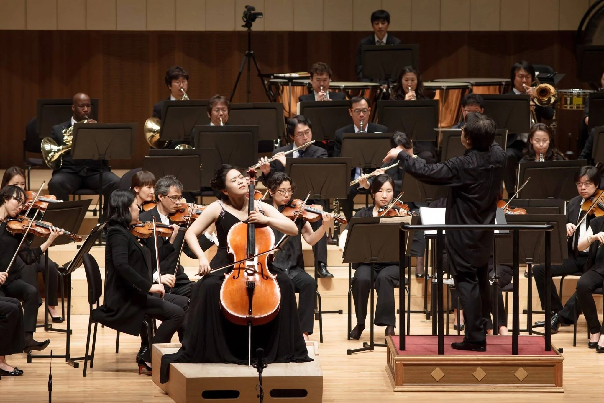 An orchestra performing on stage with a conductor directing the musicians. The stage has wooden flooring, and the musicians are seated with music stands, playing various instruments including violins, cellos, flutes, and a French horn.