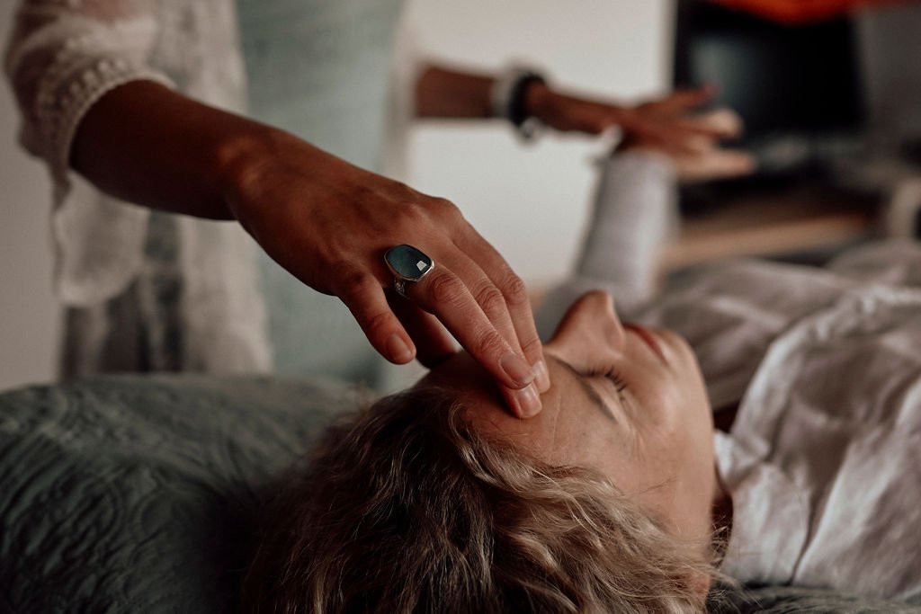 A person placing a hand on an older woman's forehead while she lies on a treatment table, suggesting a kinesiology balance.
