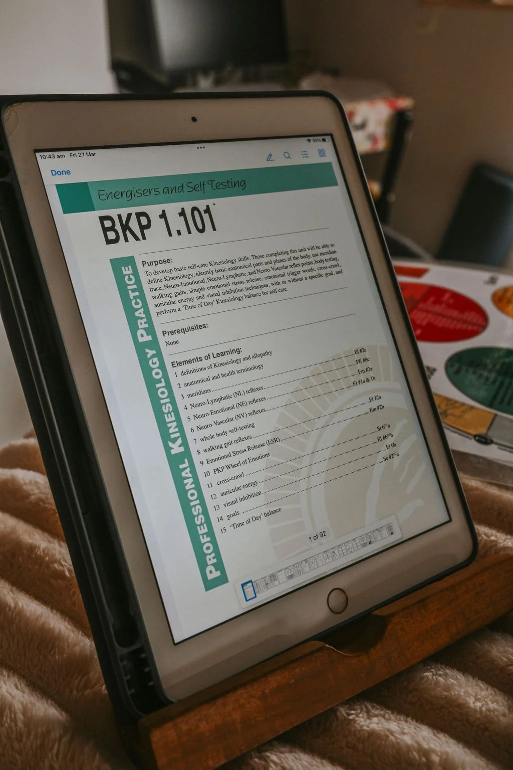 iPad displaying a document titled "BKP 1.101" with content related to Energizers and Self Testing, including sections on purpose, prerequisites, and elements of learning. The iPad is resting on a wooden stand, on a soft, brown, furry surface, with a blurred background of a room containing a coffee table with colorful items and a dark-colored chair.